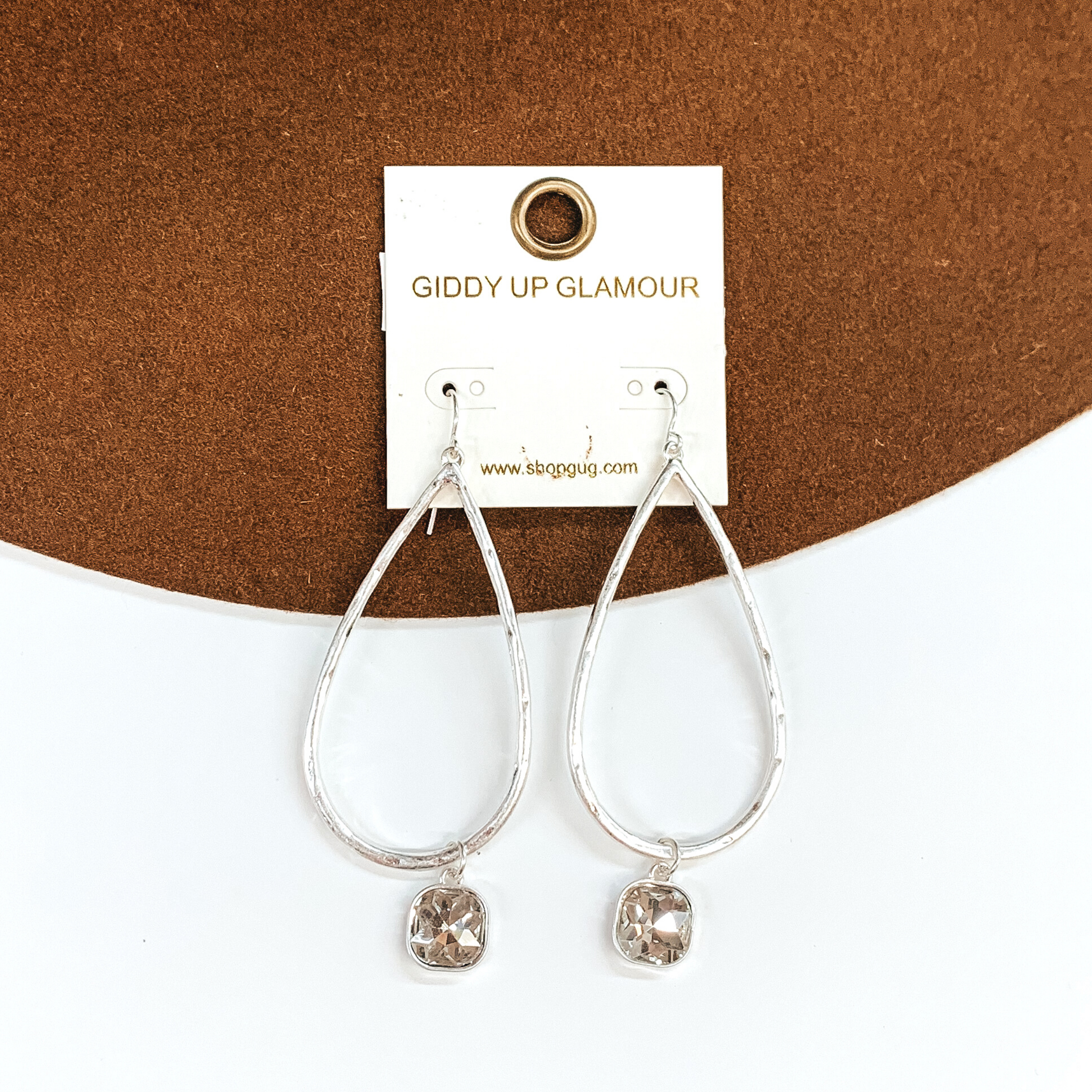 Silver teardrop hammered earrings with a hanging cushion cut clear crystal. These earrings are pictured on a white and brown background.