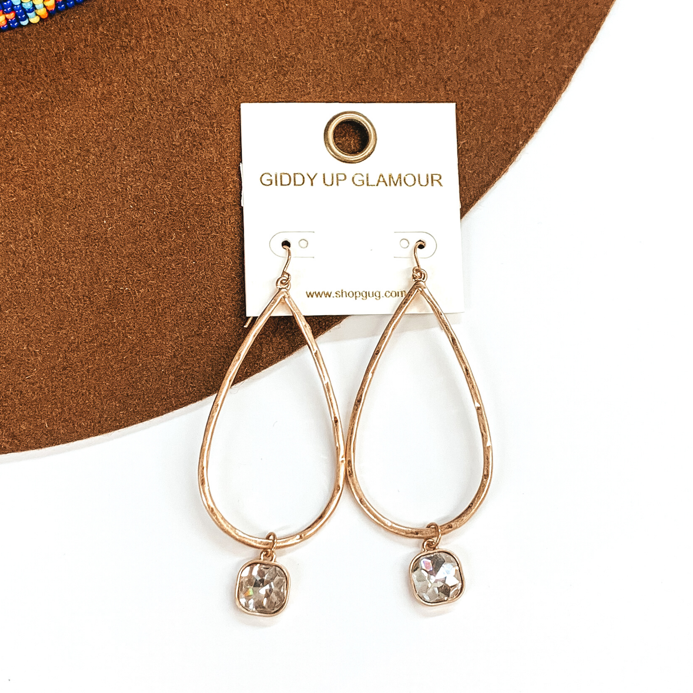 Gold teardrop hammered earrings with a hanging cushion cut clear crystal. These earrings are pictured on a white and brown background. 