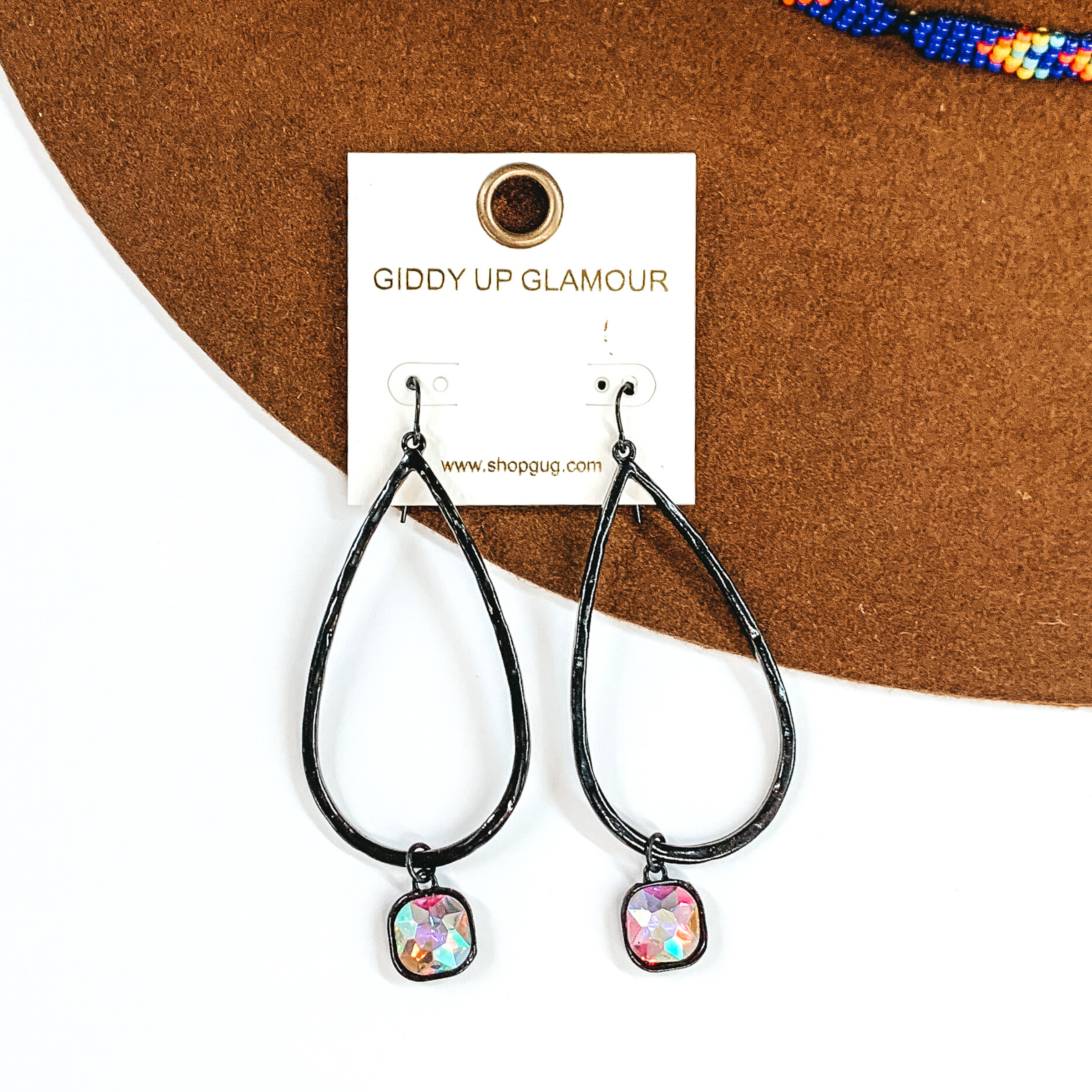 Black teardrop hammered earrings with a hanging cushion cut ab crystal. These earrings are pictured on a white and brown background.