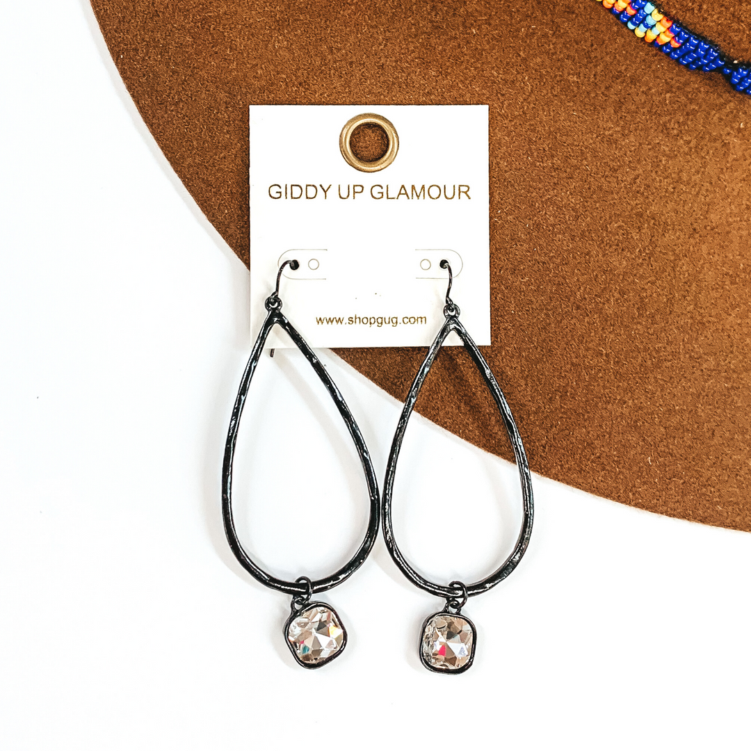 Black teardrop hammered earrings with a hanging cushion cut clear crystal. These earrings are pictured on a white and brown background.