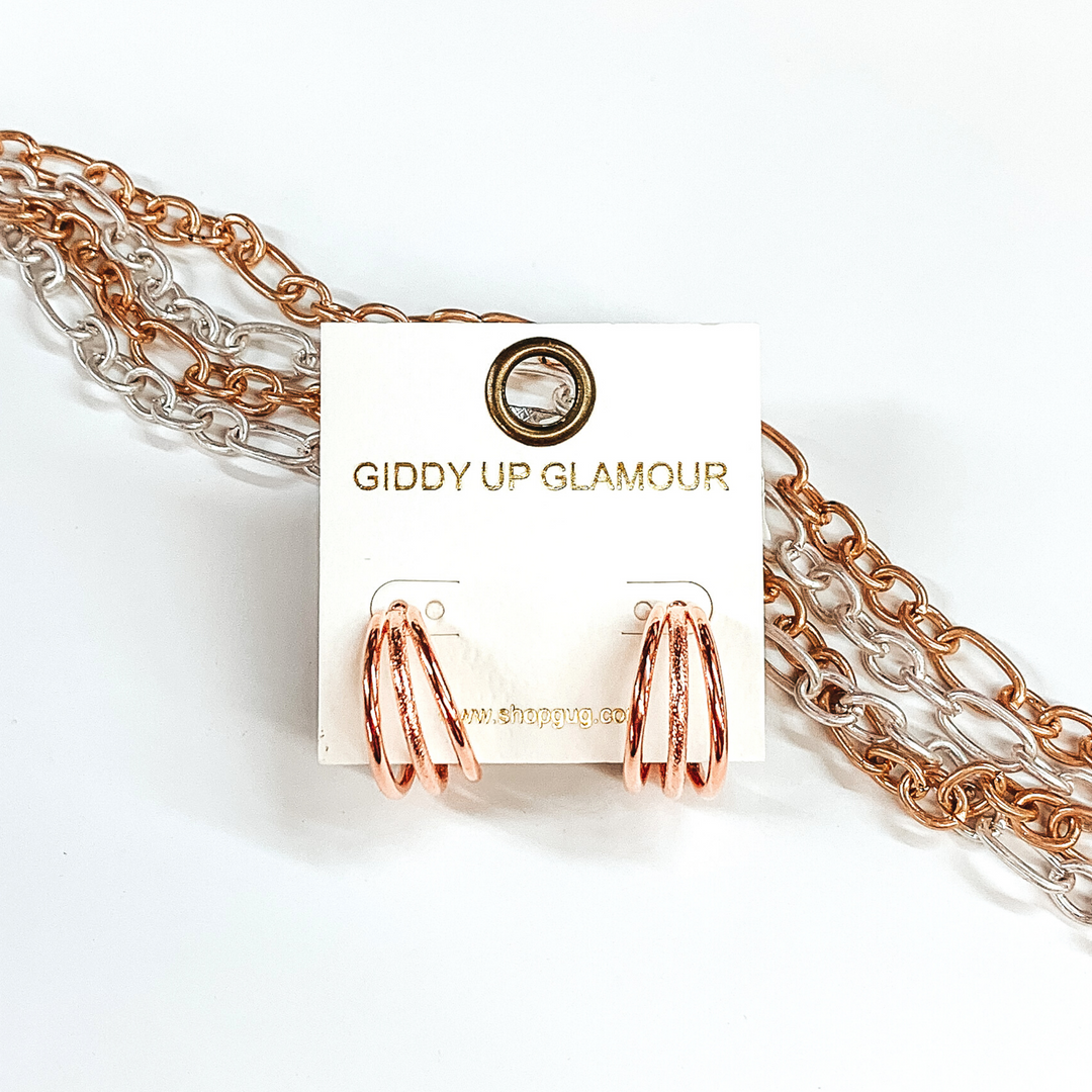 Rose gold triple circle hoop earrings. These earrings are pictured on an earring back laying on some gold and silver chains on a white background.