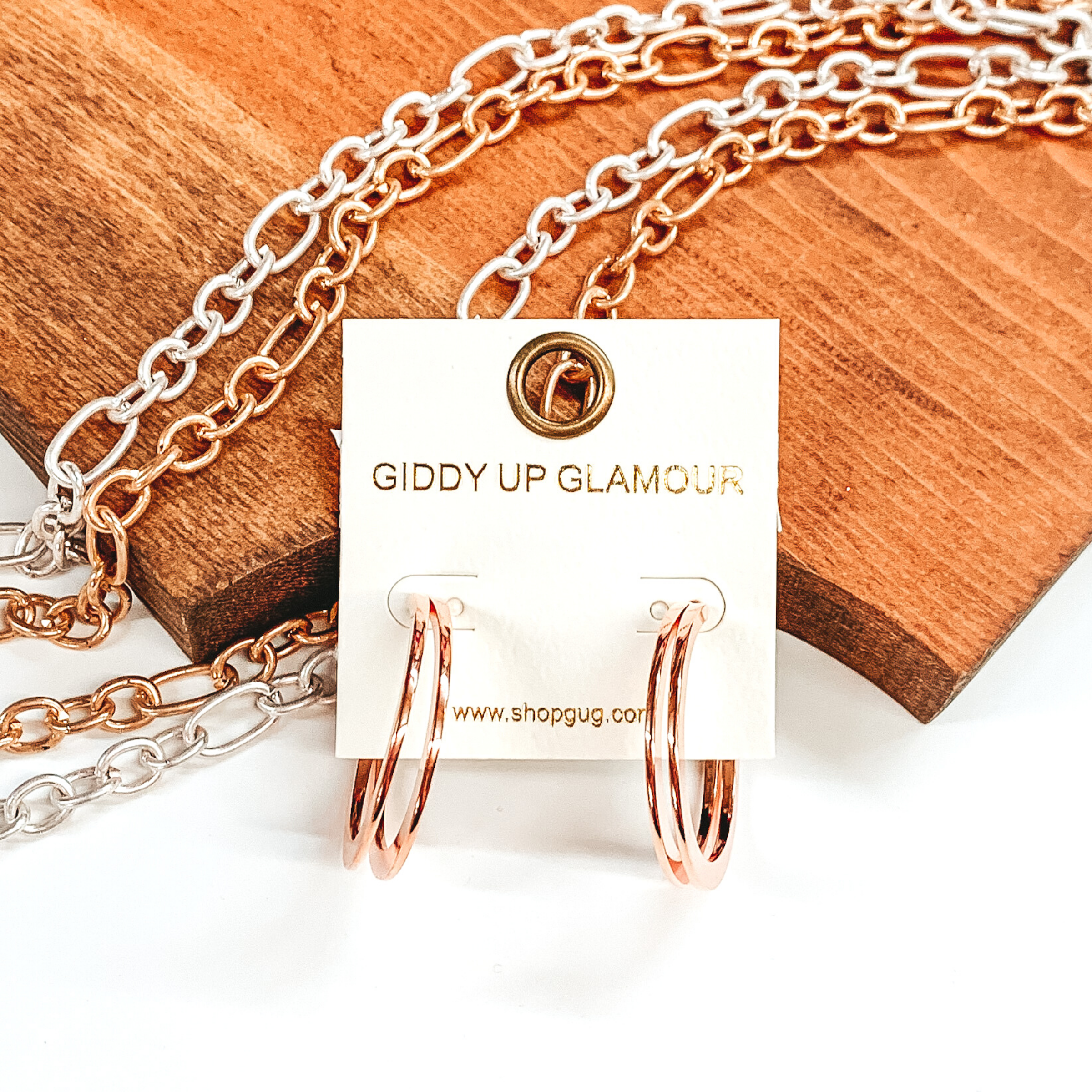 Rose gold colored circle hoops. These hoops have two circles side by side to make a double hoop. These earrings are in front of a piece of wood with silver and gold chains on it pictured on a white background. 