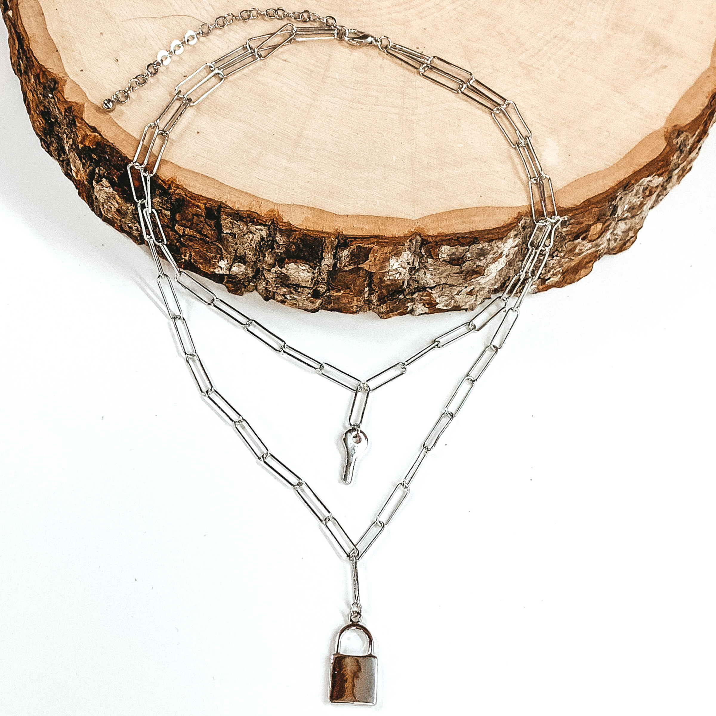Silver double chain linked necklace. the shorter strand has a key charm and the longer strand has a lock charm. This necklace is pictured laying partially on a piece of wood on a white background. 