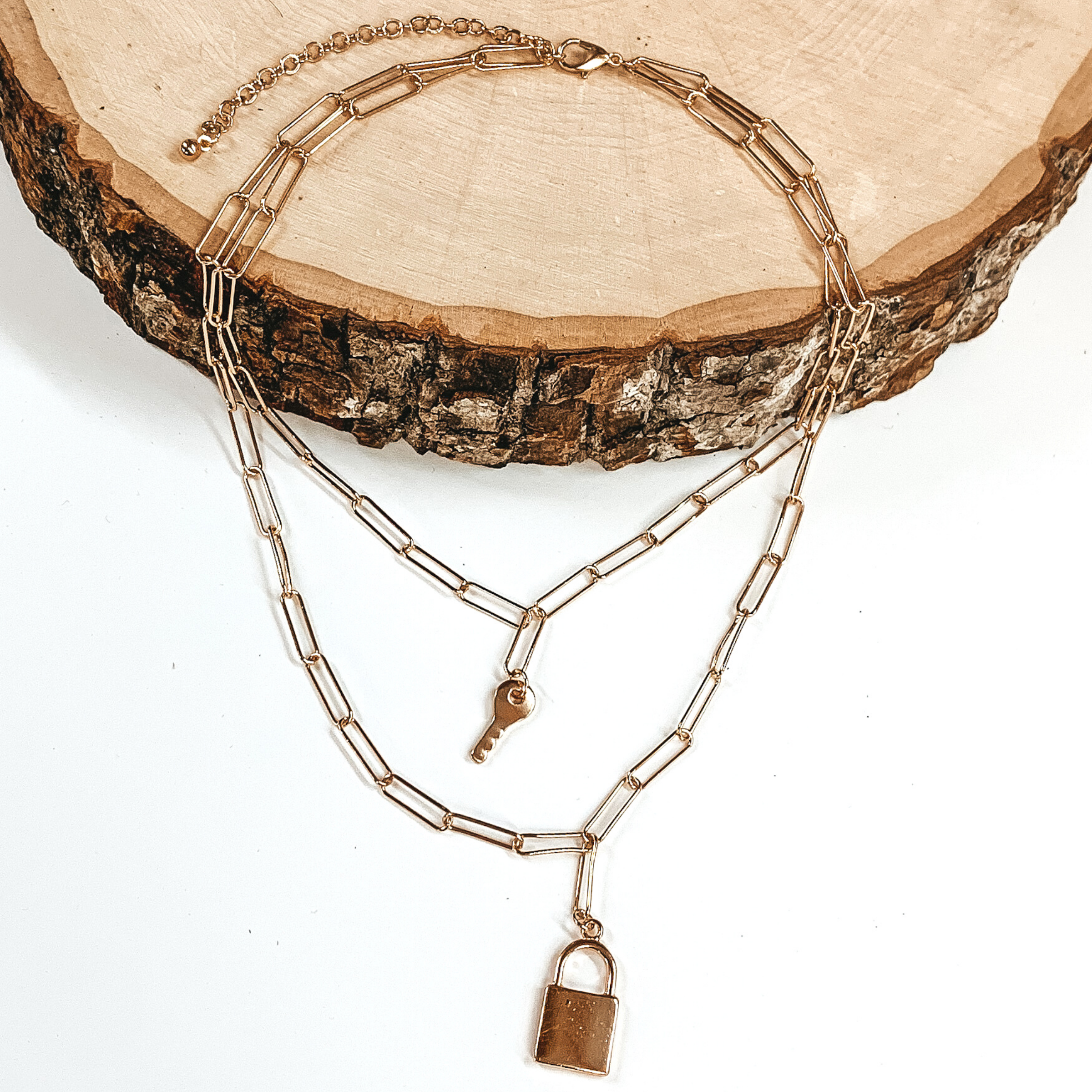 Gold double chain linked necklace. the shorter strand has a key charm and the longer strand has a lock charm. This necklace is pictured laying partially on a piece of wood on a white background.
