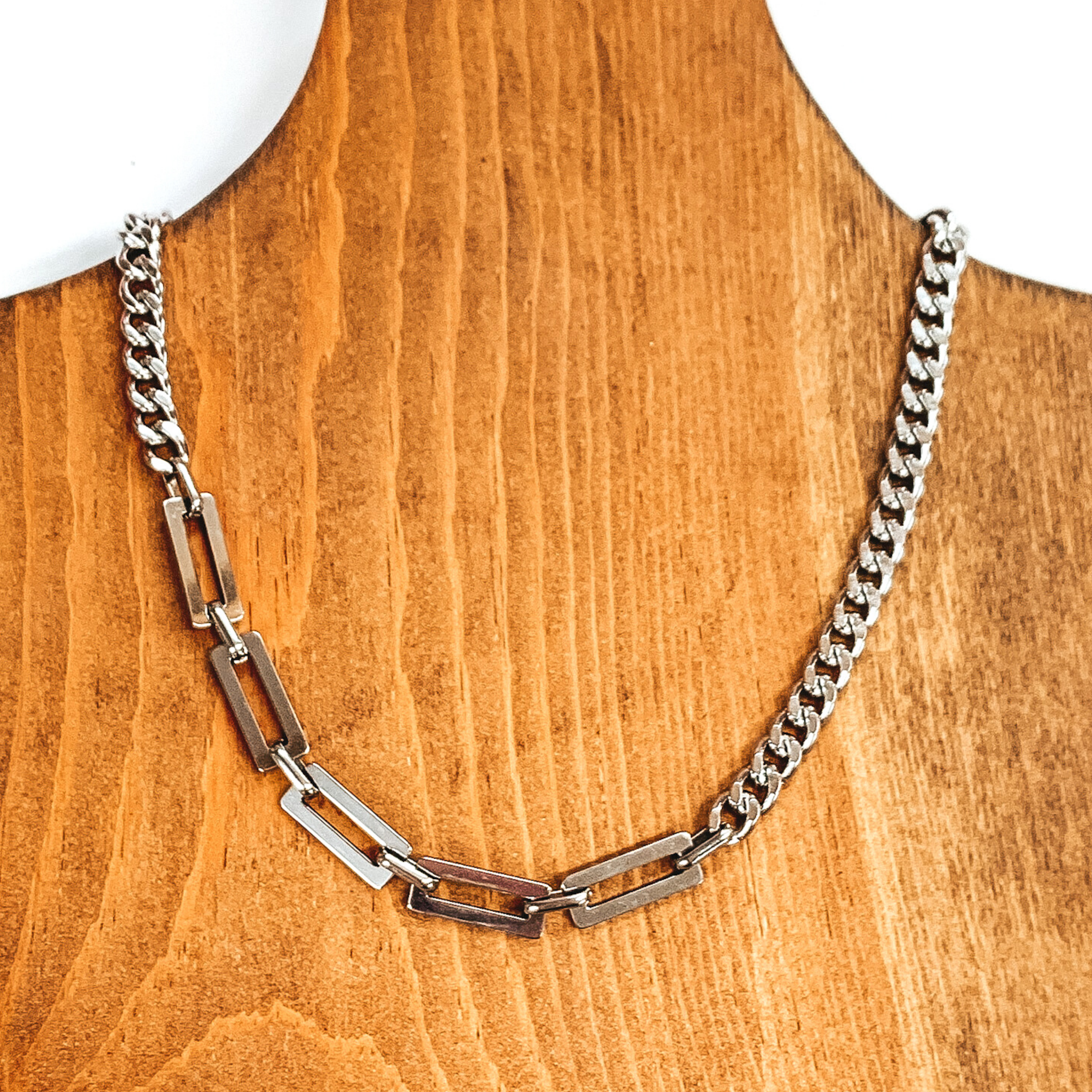 Silver multi chained necklace laying on a wood necklace holder. This necklace is mostly a curb chain and there is a small segment that has thick rectangle link part. This is all pictured on a white background. 
