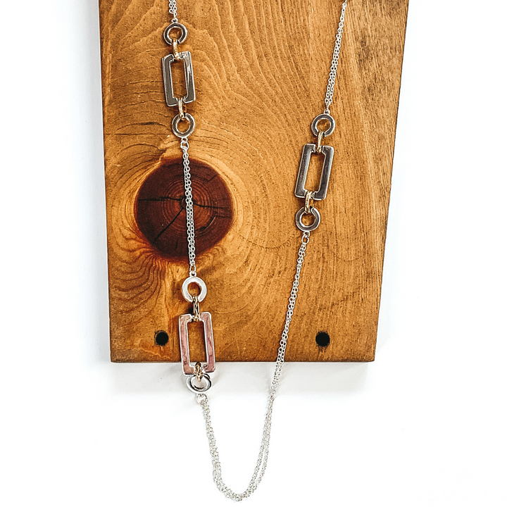 Silver necklace with multi shape spacers. In each spacer there is a rectangle center link, then there is a thick gold link on either side, and finally it has a circle silver link on each end. this necklace is pictured laying on a dark piece of wood on a white background. 