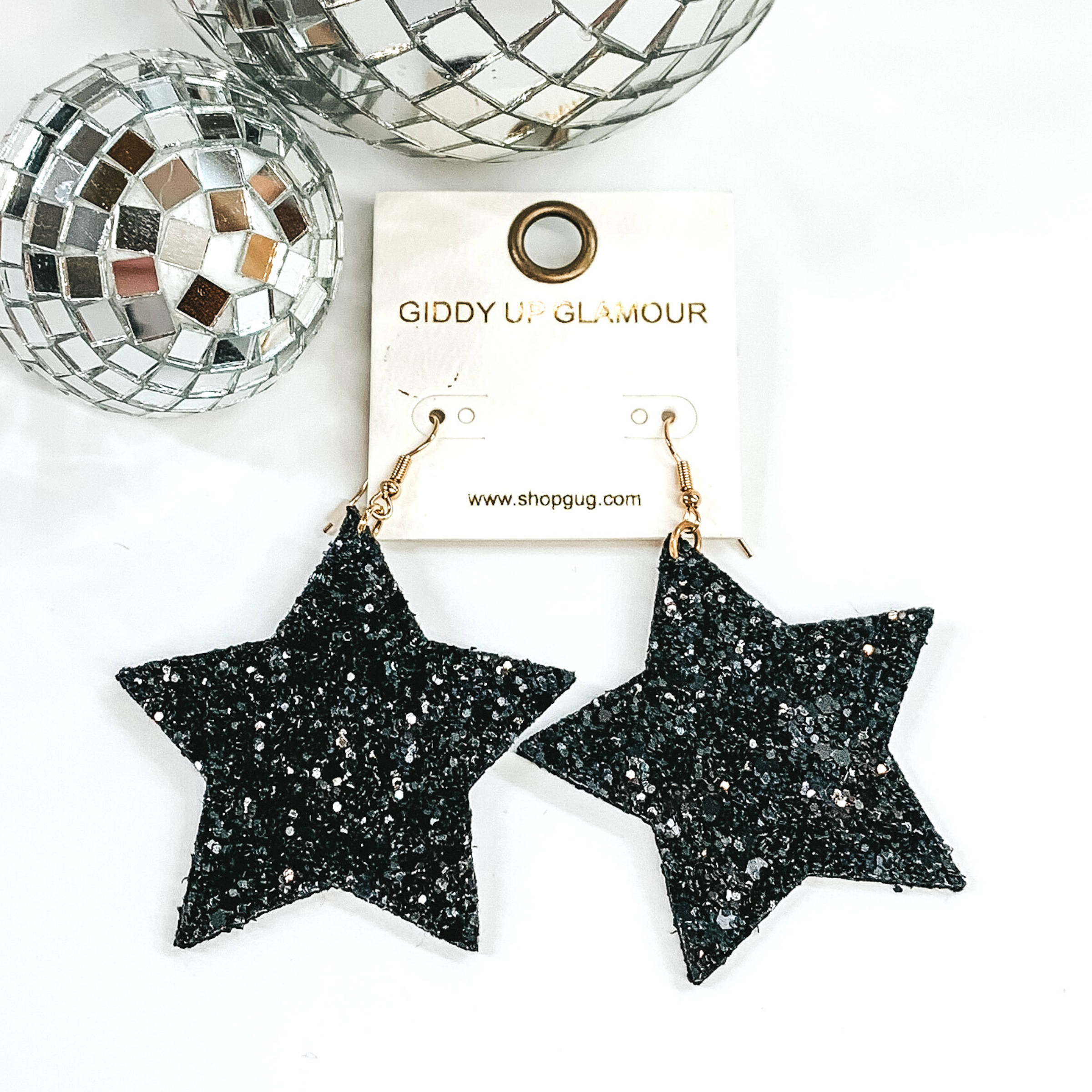 Glitter star dangle earrings in black with a gold earrings hook. These earrings are pictured on a white background with a disco ball at the top of the picture. 