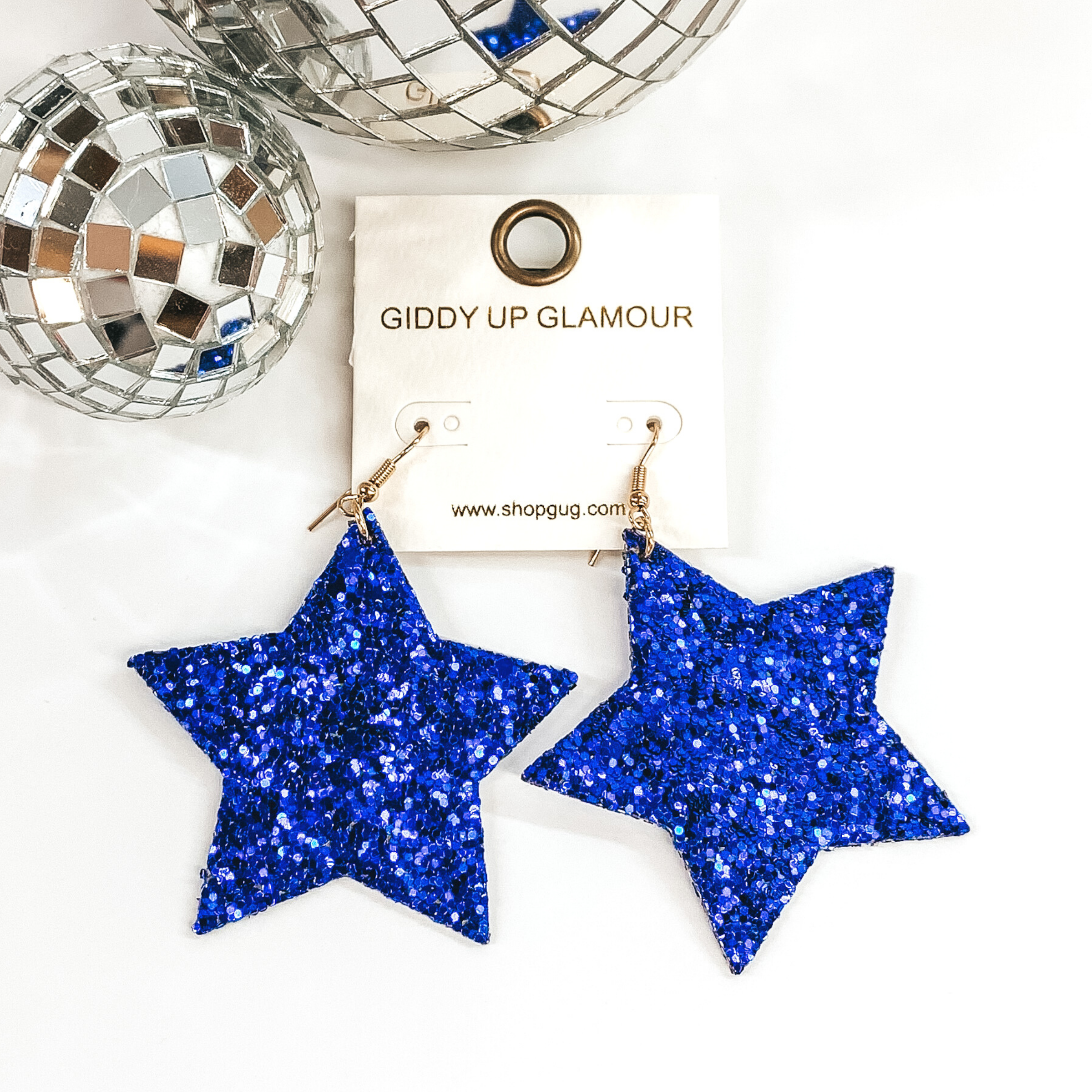 Glitter star dangle earrings in blue with a gold earrings hook. These earrings are pictured on a white background with a disco ball at the top of the picture.