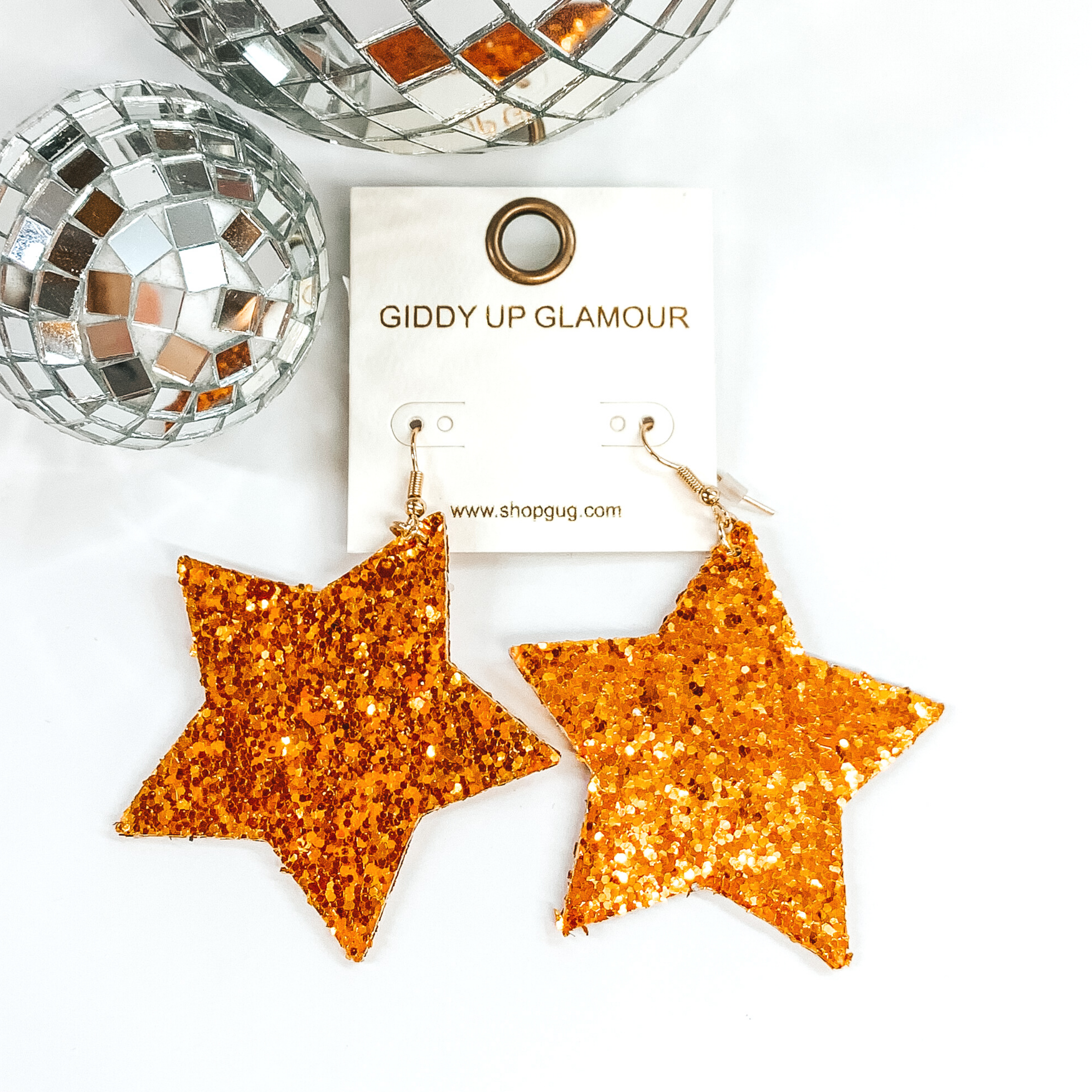 Glitter star dangle earrings in orange with a gold earrings hook. These earrings are pictured on a white background with a disco ball at the top of the picture.