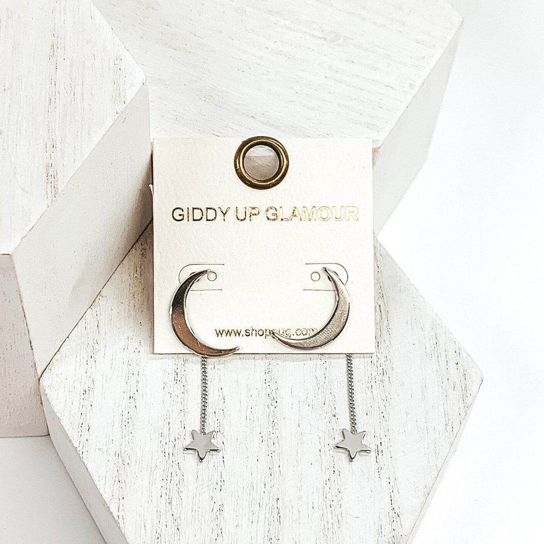 Silver moon earrings with a hanging star charm at the bottom of each earring. These earrings are pictured on a white earring holder and is laying on white blocks on a white background.