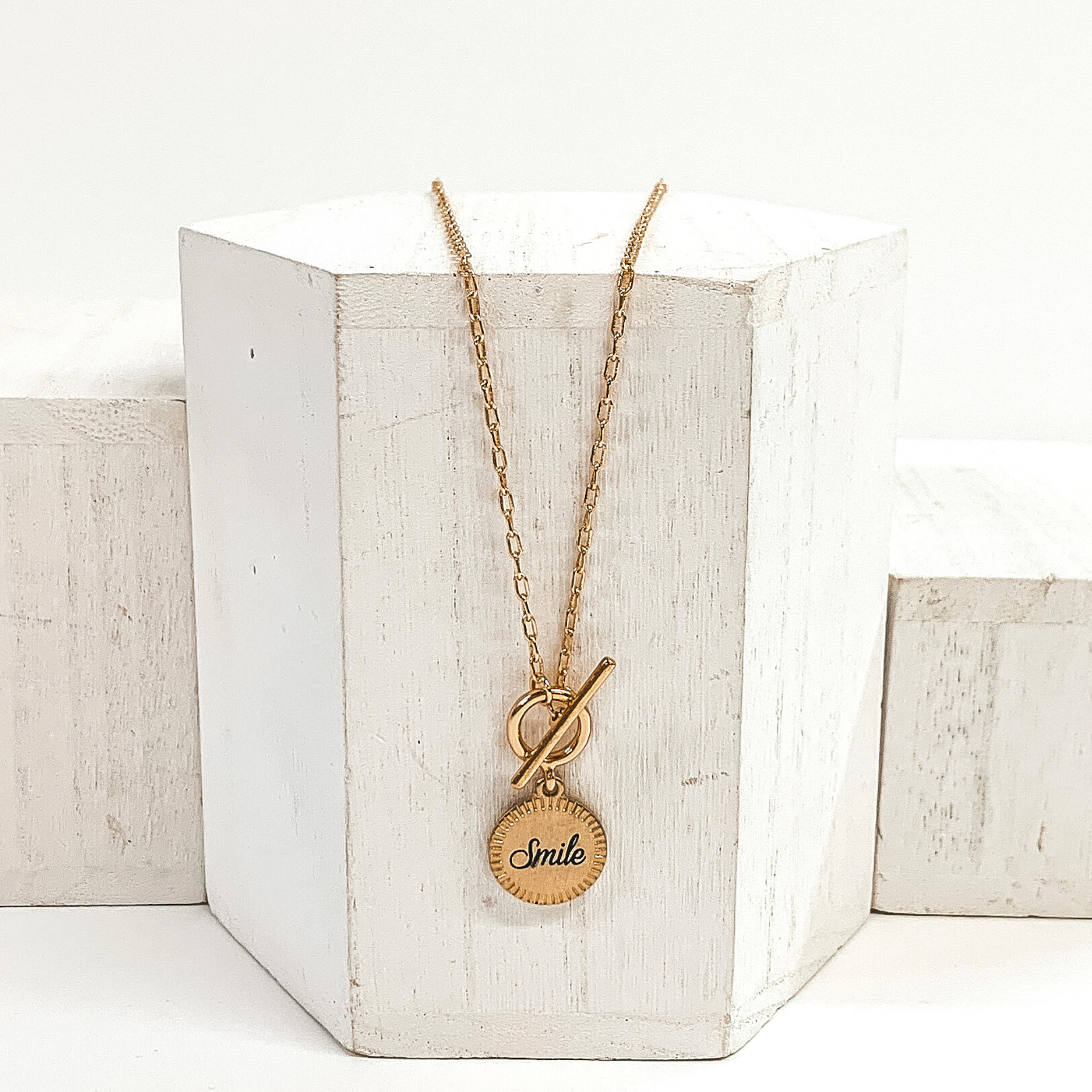 This is a gold necklace that has a front toggle clasp and a circle pendant with the word "smile" written in the center. This necklace is pictured laying on a white block and on a white background.