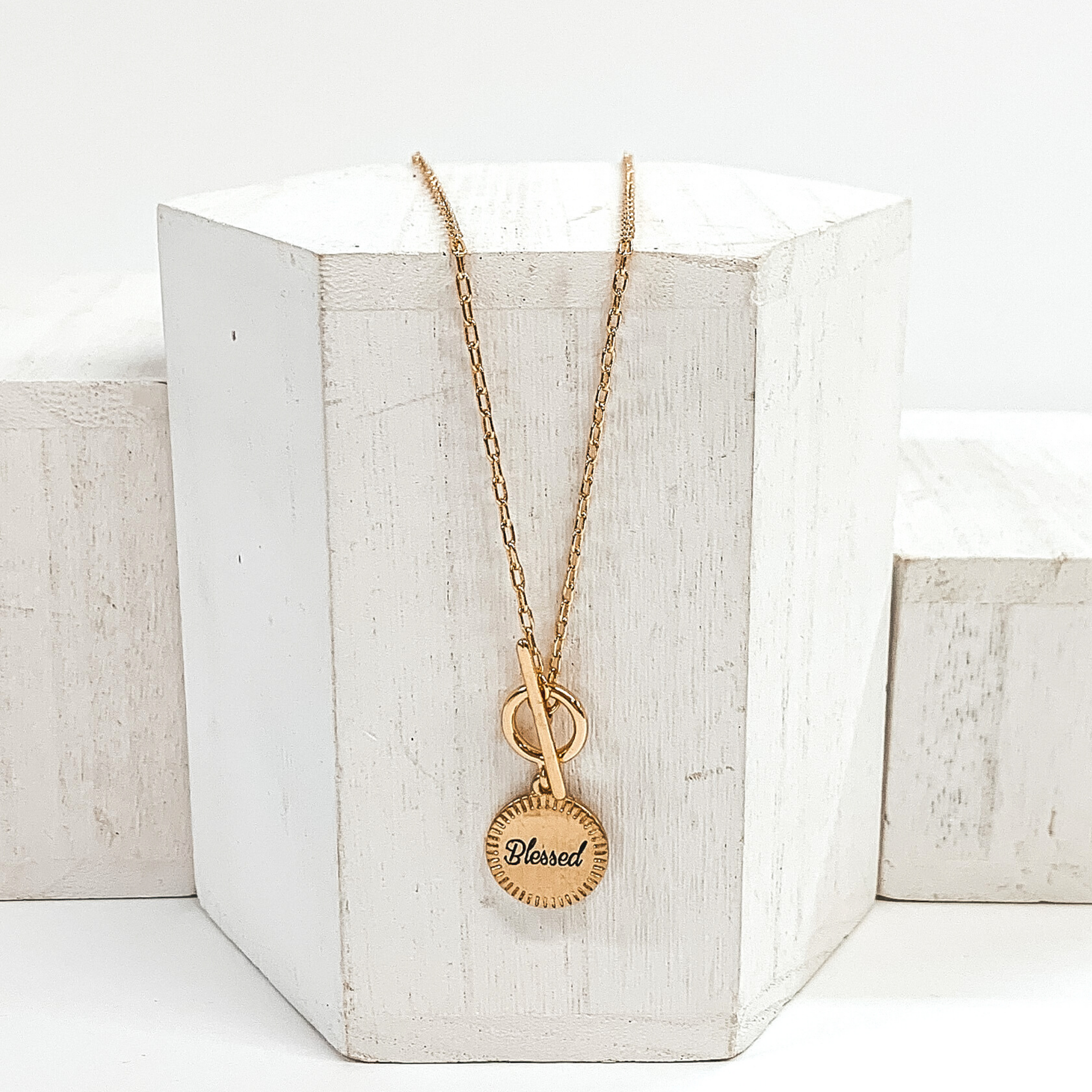 This is a gold necklace that has a front toggle clasp and a circle pendant with the word "blessed" written in the center. This neecklace is pictured laying on a white block and on a white background.