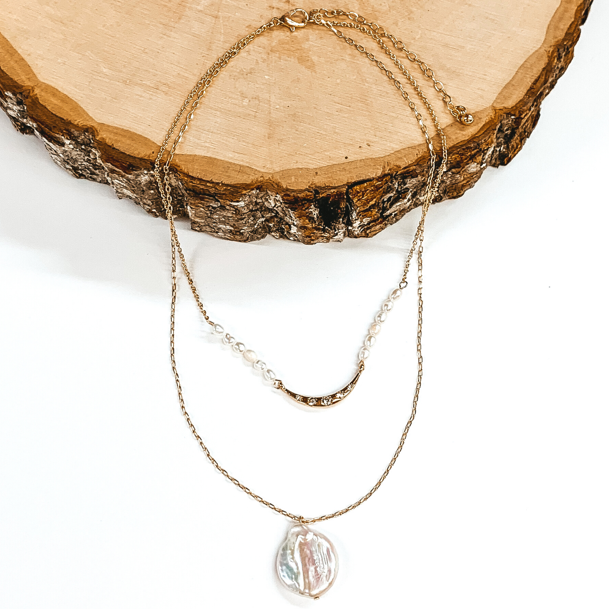 Gold double layered necklace. The shorter chain has a middle gold bar with white beads on either side of the bar. The longer strand has a single white pendant. This necklace is pictured laying partially on a piece of wood on a white background.