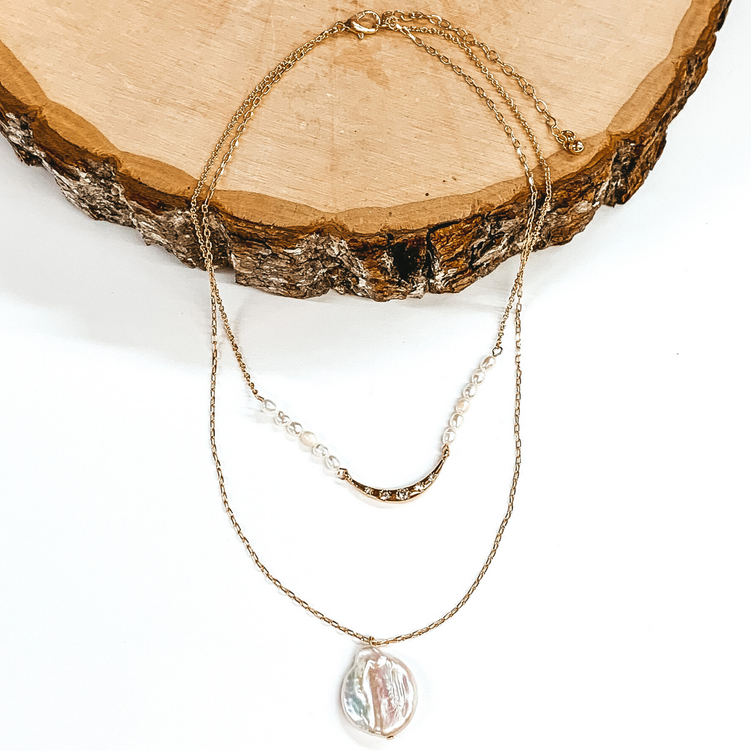Gold double layered necklace. The shorter chain has a middle gold bar with white beads on either side of the bar. The longer strand has a single white pendant. This necklace is pictured laying partially on a piece of wood on a white background.