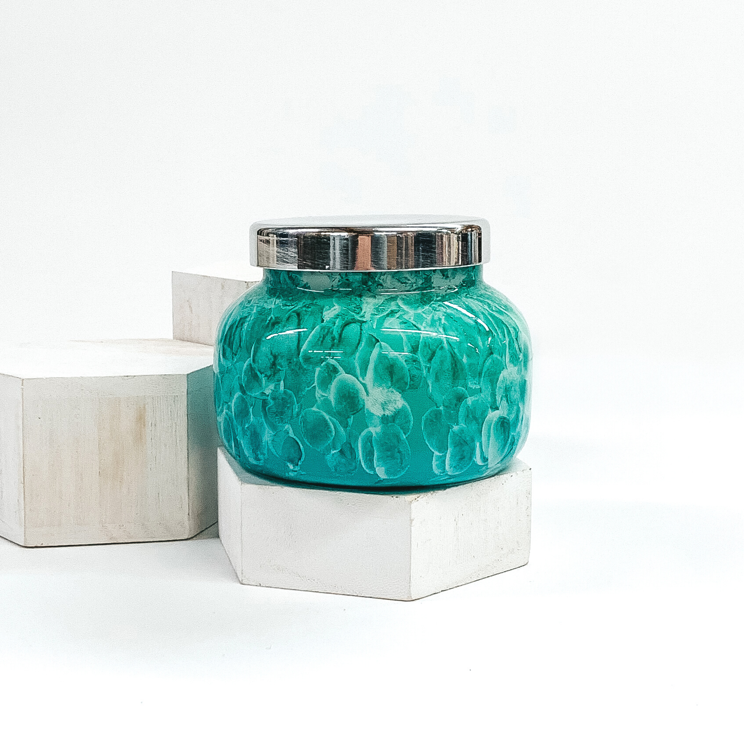 A candle in a watercolor mint jar with a silver lid. The candle is pictured sitting on a white block with two other blocks behind it. All pictured on a white background. 