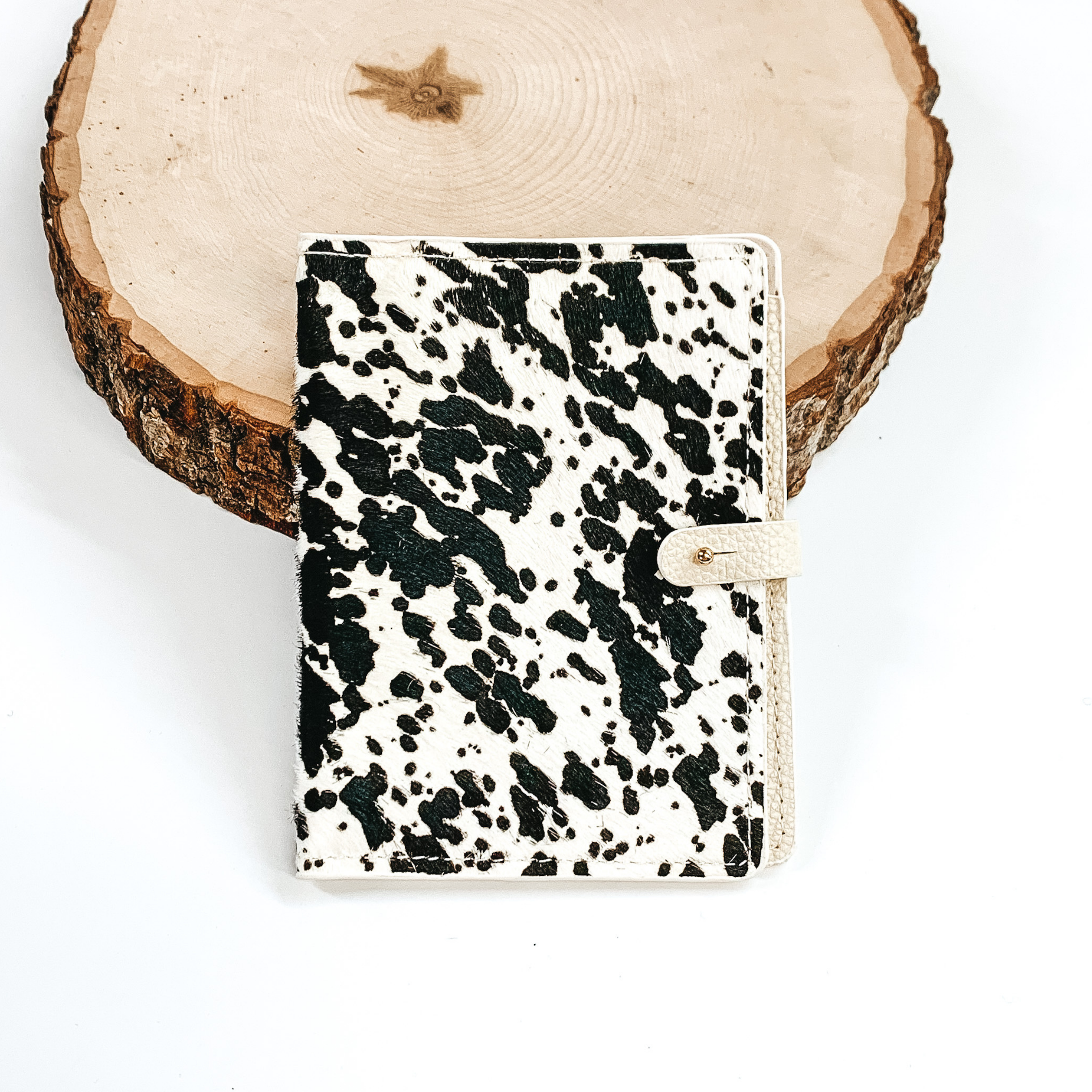 White and black cow print wallet passport with a gold button to close. This wallet is pictured laying against a piece of wood on a white background. 