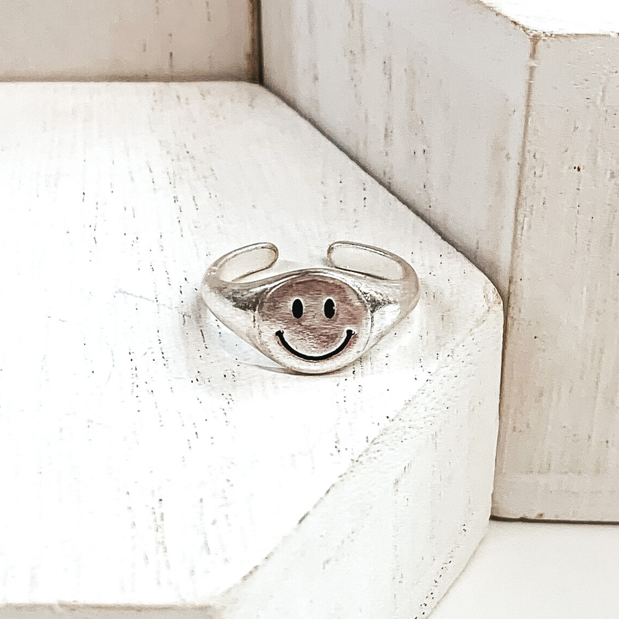 Silver, non adjustable ring with a round smiley face. This ring is pictured on a white block with more white blocks in the background. 