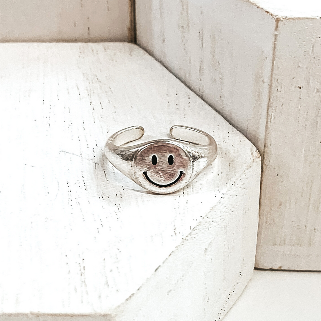 Silver, non adjustable ring with a round smiley face. This ring is pictured on a white block with more white blocks in the background. 
