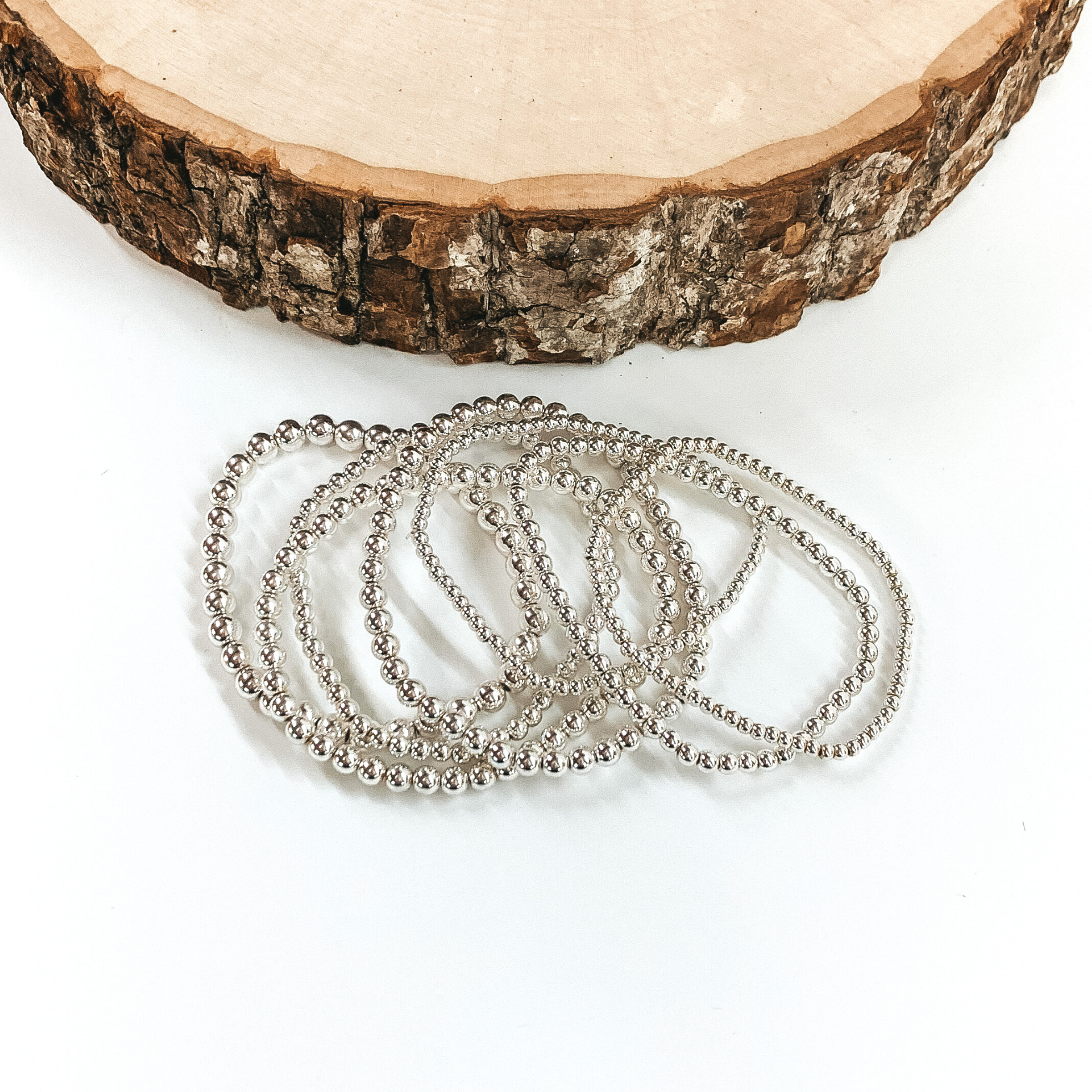 A set of seven silver beaded bracelets ranging in different size beads. These bracelets are laying on top of each other in front of a piece of wood on a white background.