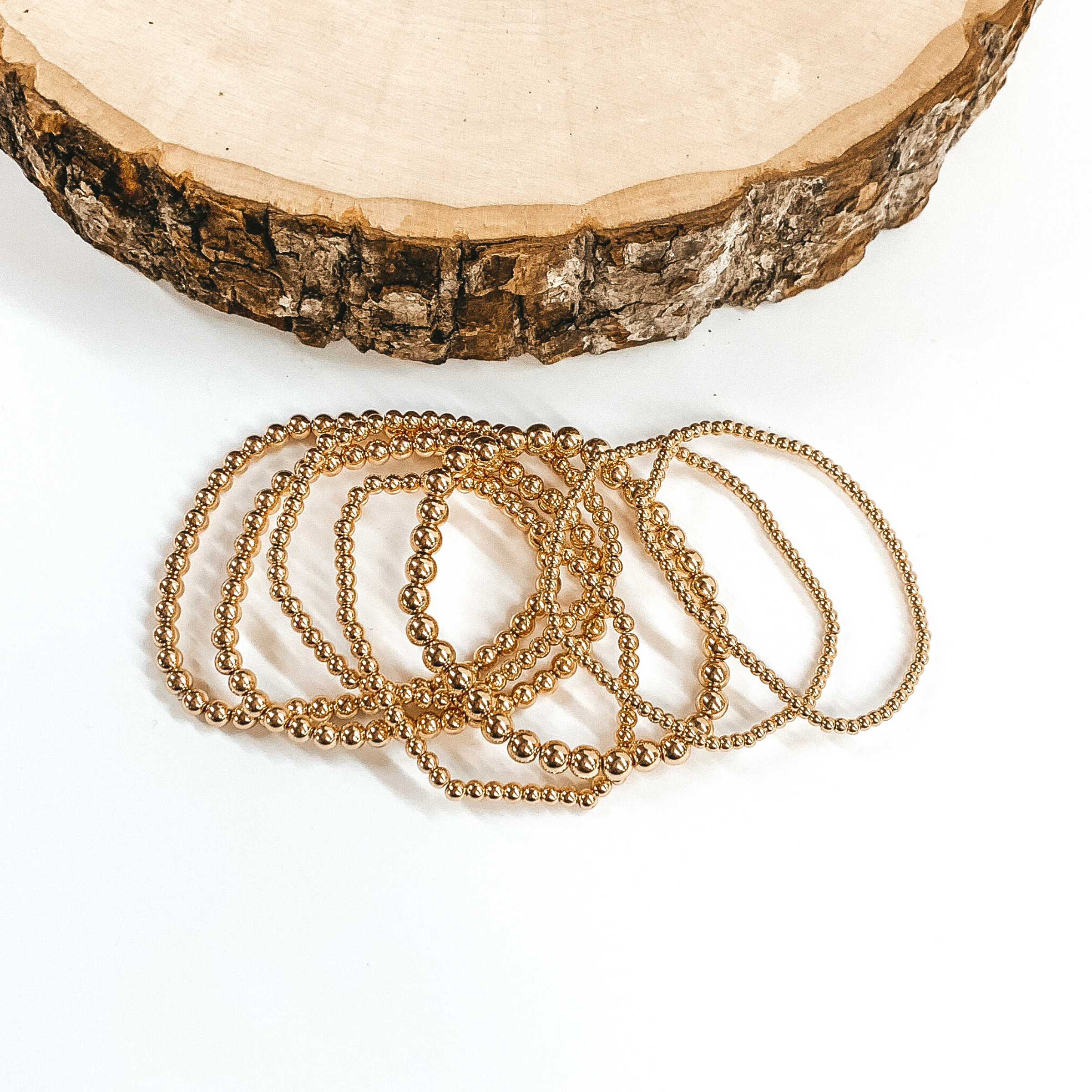 A set of seven gold beaded bracelets ranging in different size beads. These bracelets are laying on top of each other in front of a piece of wood on a white background. 