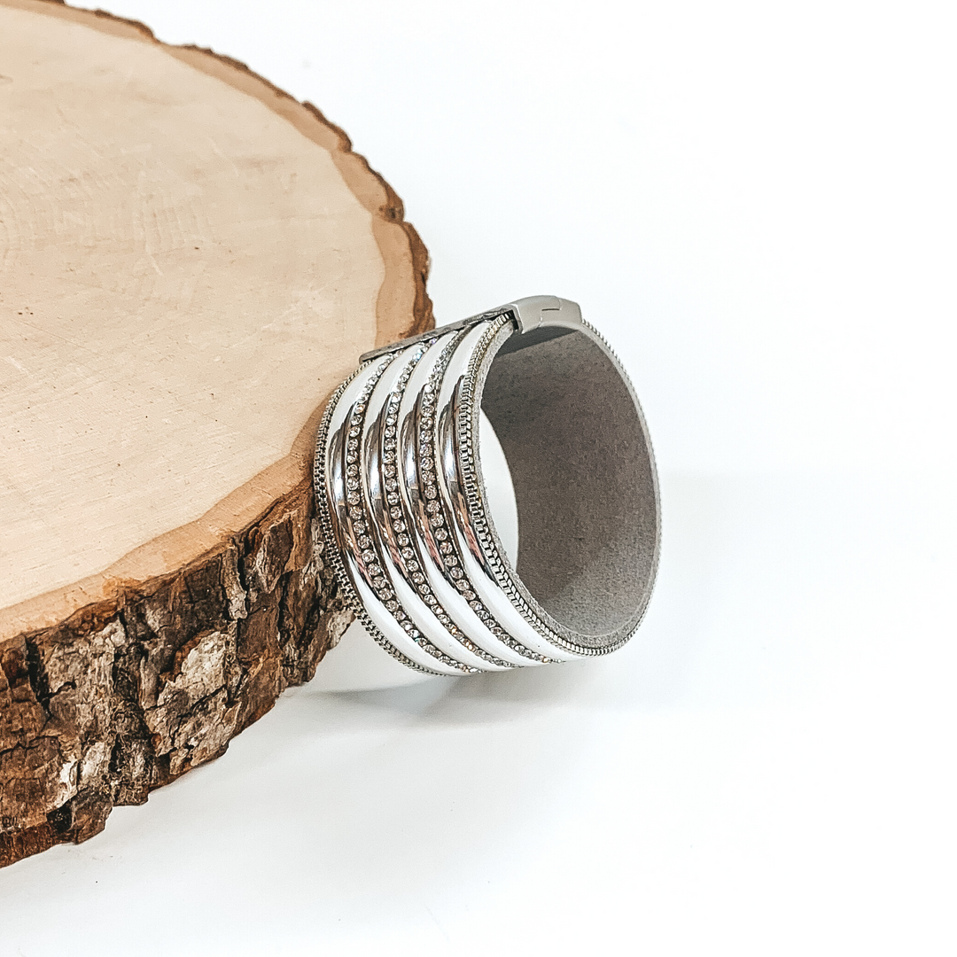 Wide band with silver colored rows separated by clear crystals. It has a silver clasp. This bracelet is pictured laying against a piece of wood on a white background.