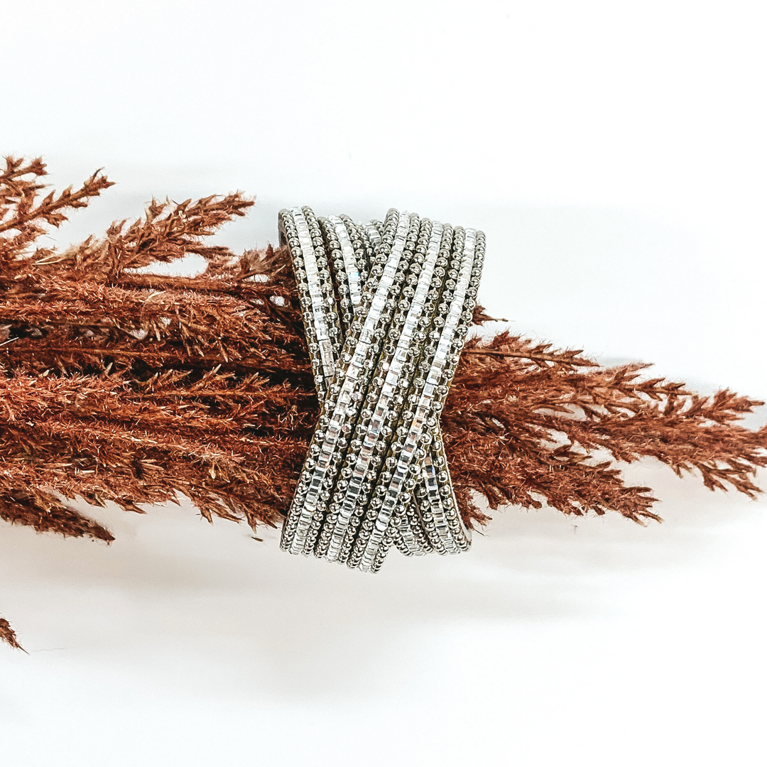 Silver strands that has clear crystals in the middle of each strand. Two sections of three strands each are crossed over each other in the middle. This bracelet is pictured on a piece of brown floral on a white background.