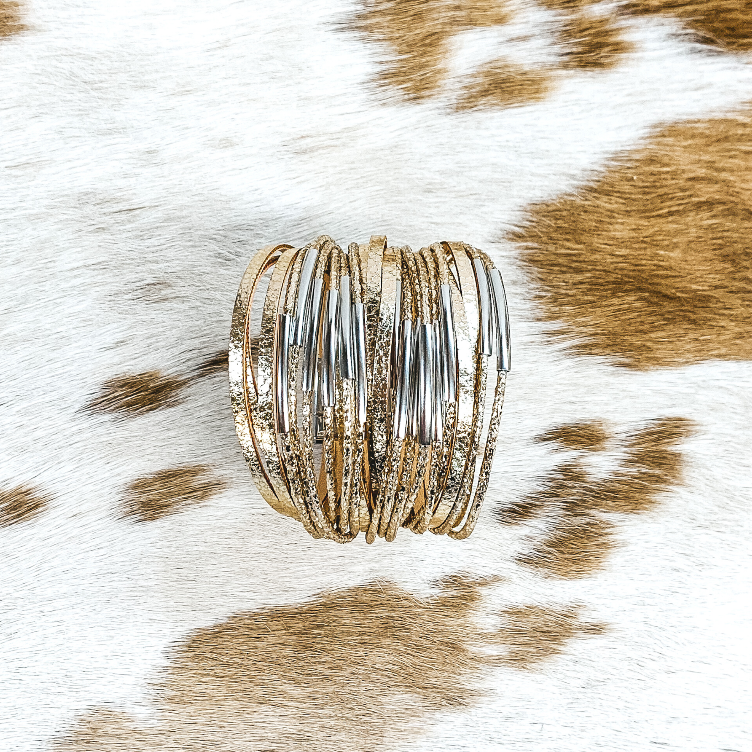 This bracelet has a lot of gold destressed strands with some of them having silver bar pendants. This bracelet is pictured on a white and brown cow print background. 
