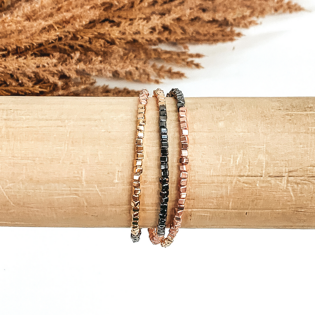 Three stranded square beaded bracelet. The colors included are gold, gunmetal, and rose gold. This bracelet is pictured on a wood bracelet holder on a white background with some tan floral at the top of the picture.