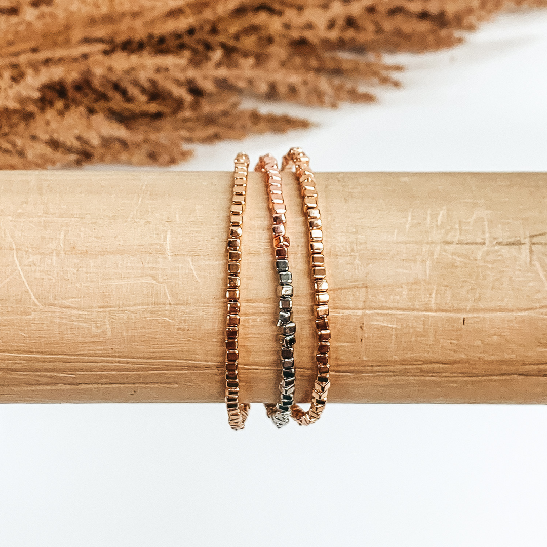 Three stranded square beaded bracelet. The colors included are gold, silver, and rose gold. This bracelet is pictured on a wood bracelet holder on a white background with some tan floral at the top of the picture. 
