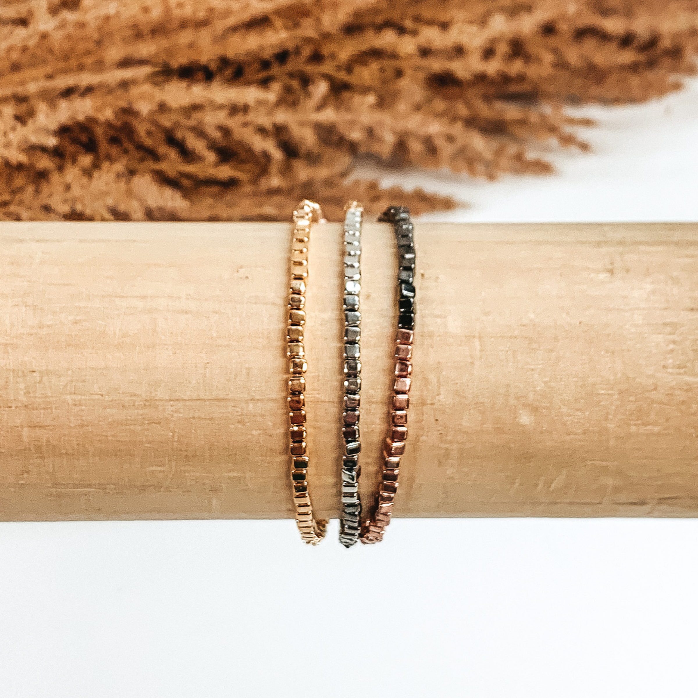Three stranded square beaded bracelet. The colors included are gold, silver, gunmetal, and rose gold. This bracelet is pictured on a wood bracelet holder on a white background with some tan floral at the top of the picture. 