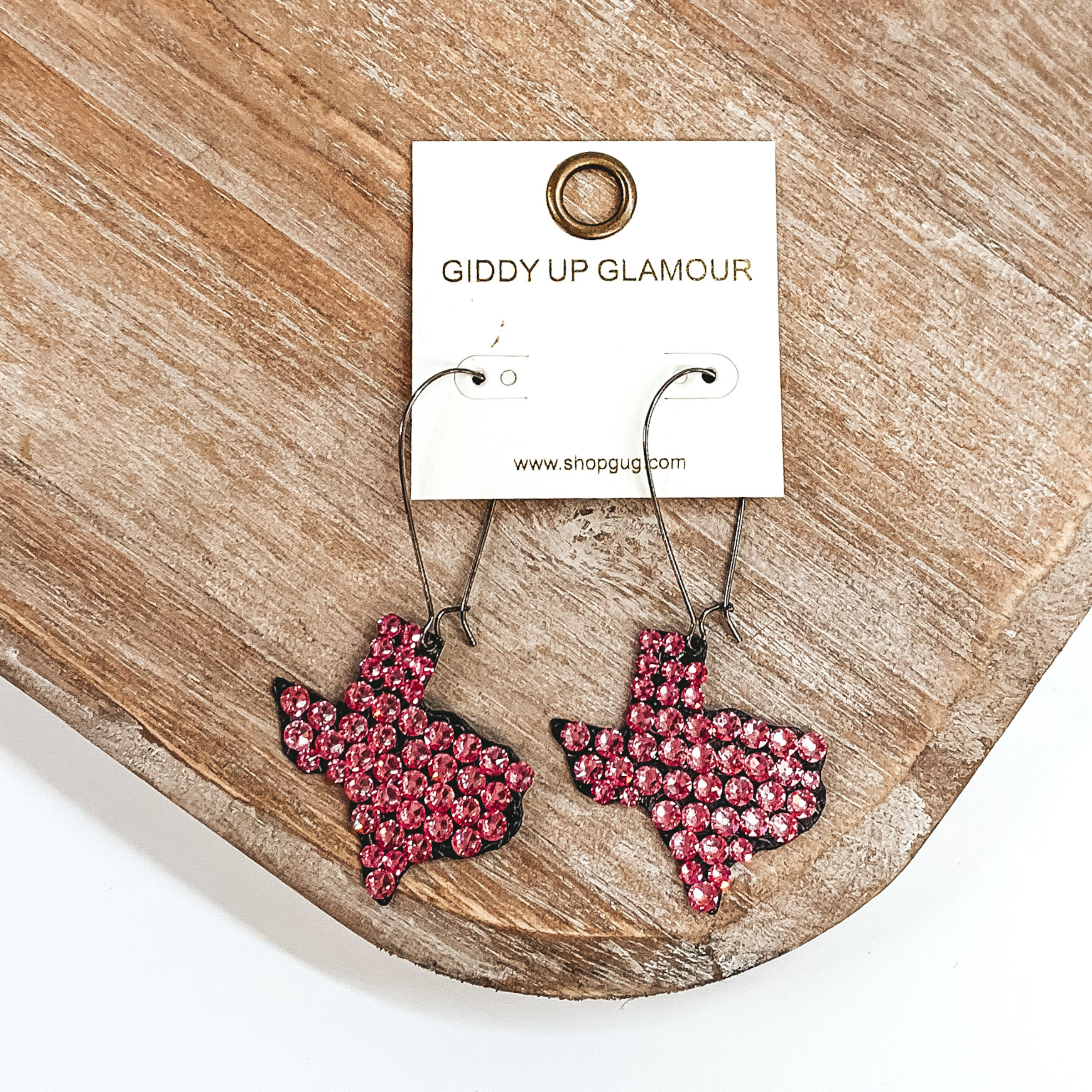 Black thin wire earringd with a texas shaped pendant. the pendant is covered in pink crystals. These earrings are pictured on a tan wood colored prop on a white background. 