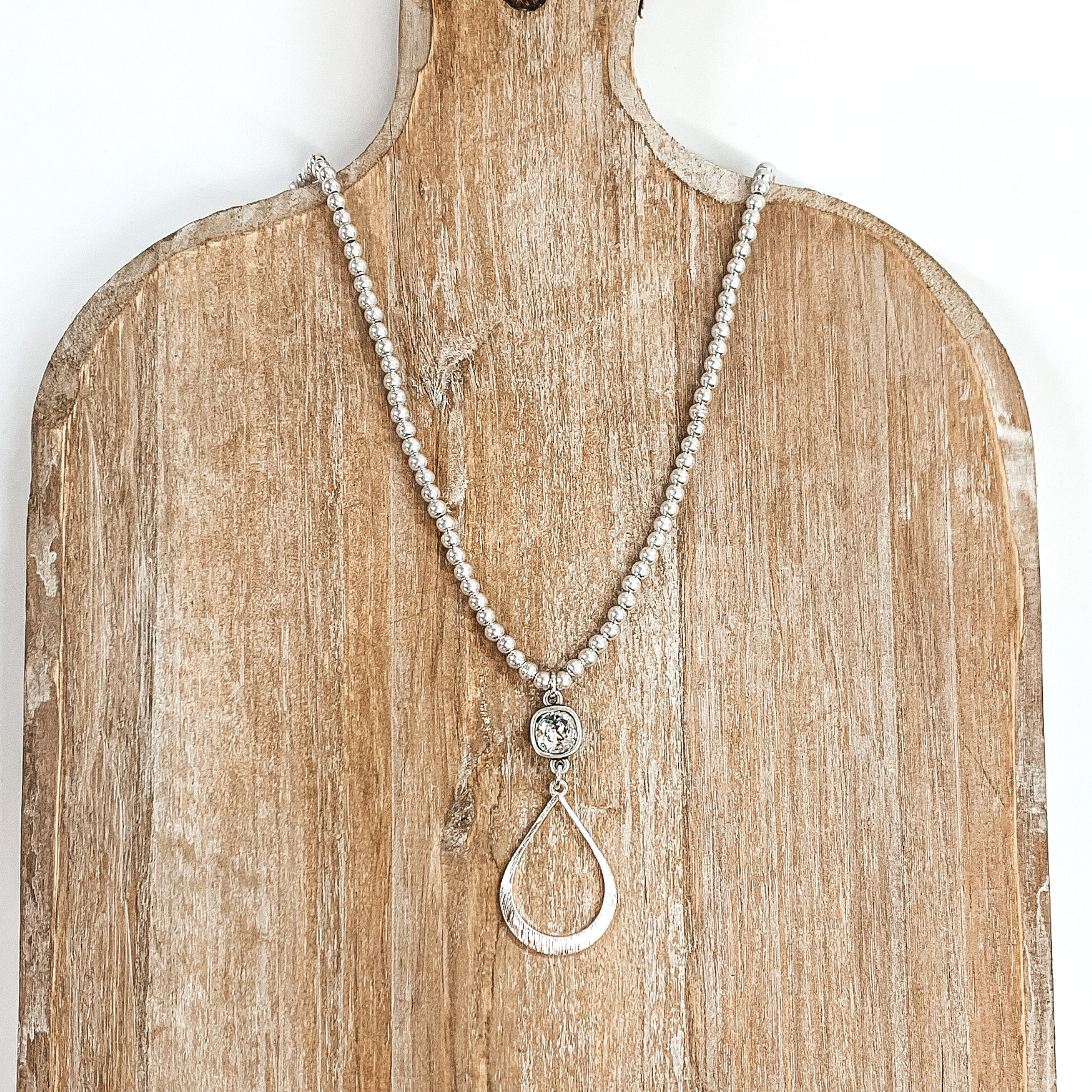 Silver beaded necklace with hanging cushion cut crystal and open teardrop pendant. This necklace is pictured on a tan necklace holder and on a white background. 