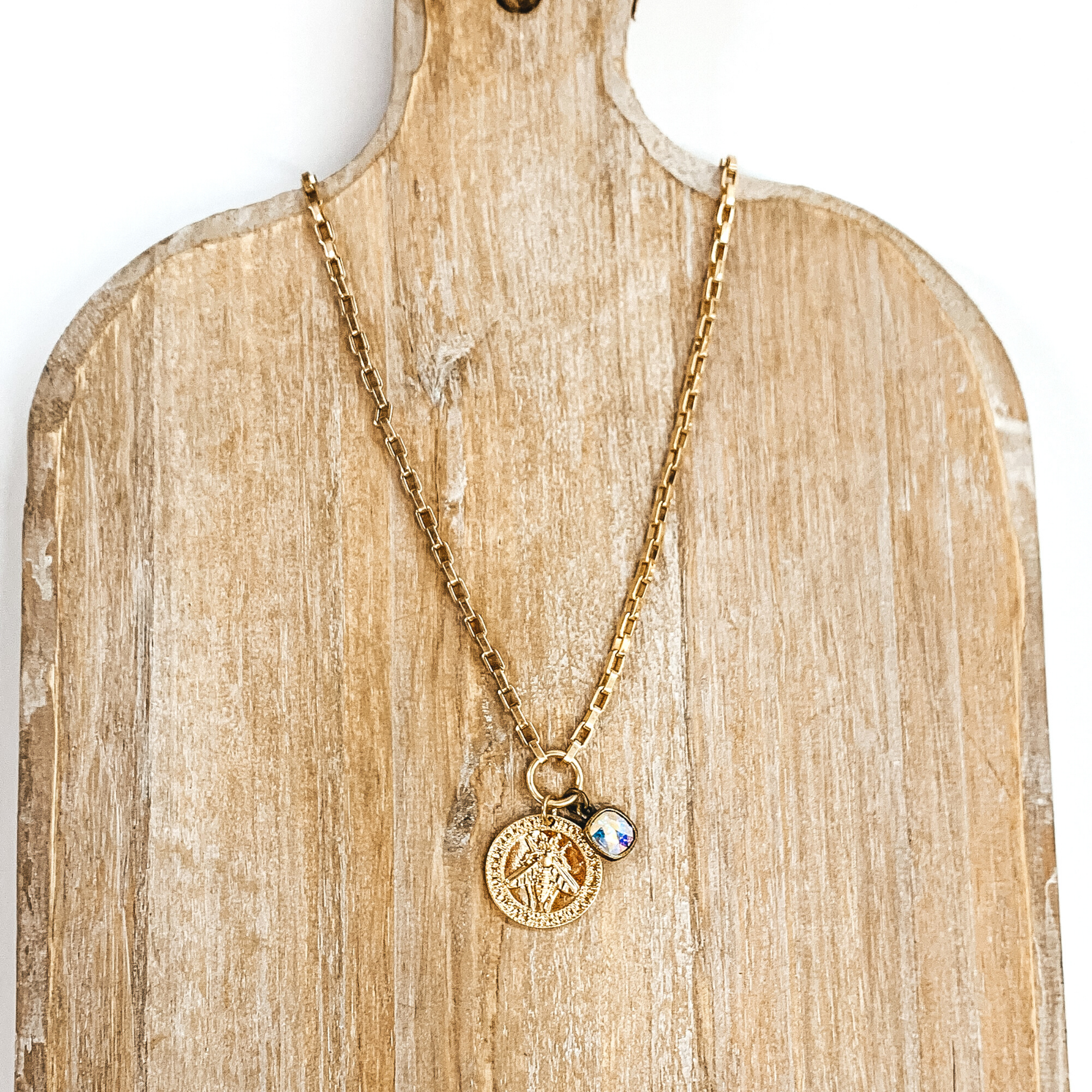 Gold chained necklace with a hanging gold coin pendant and hanging ab crystal. On the coin there is an engraved bee. This necklace is pictured on a tan necklace holder and on a white background. 