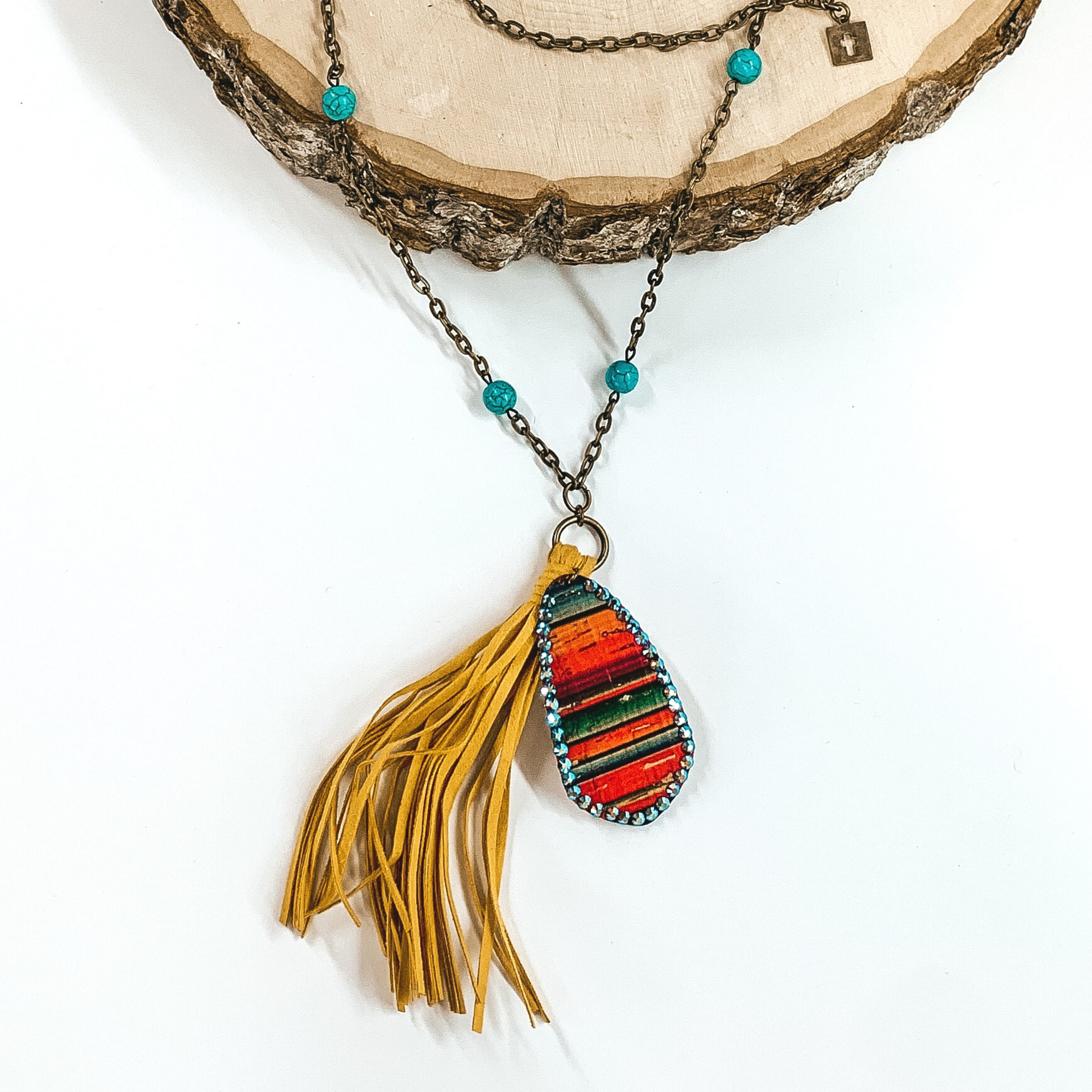 Long bronze chained necklace with turquoise bead spacer. At the bottom of the necklace there is a yellow tassel with an oval serape charm that has a crystal outline. This necklace is partially laying on a piece of wood on a white background. 