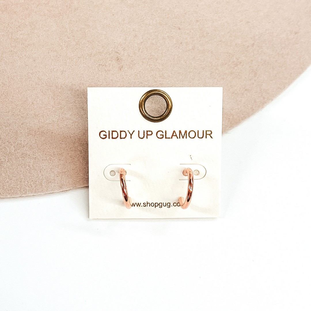 Thin rose gold hoops on a white earrings holder. These hoops are pictured on a white and beige background. 