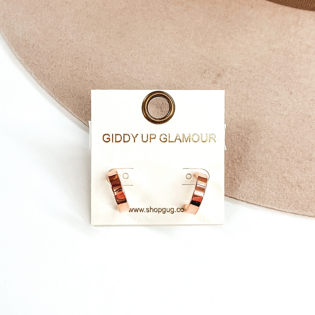 Flat rose gold hoops on a white earrings holder. These earrings are pictured on a white and beige background.