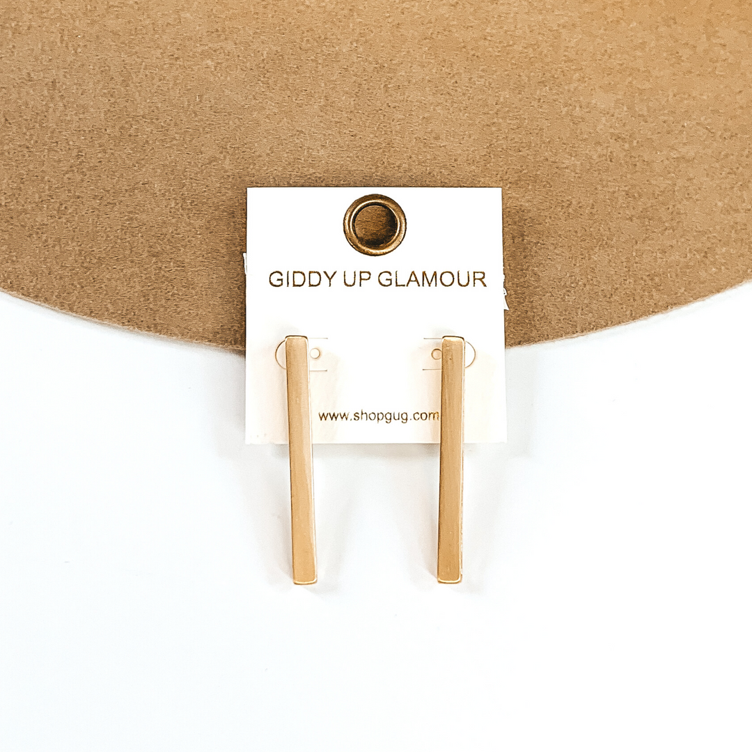Gold rectangle bar earrings on a white earrings holder. These earrings are pictured on a white and tan background. 