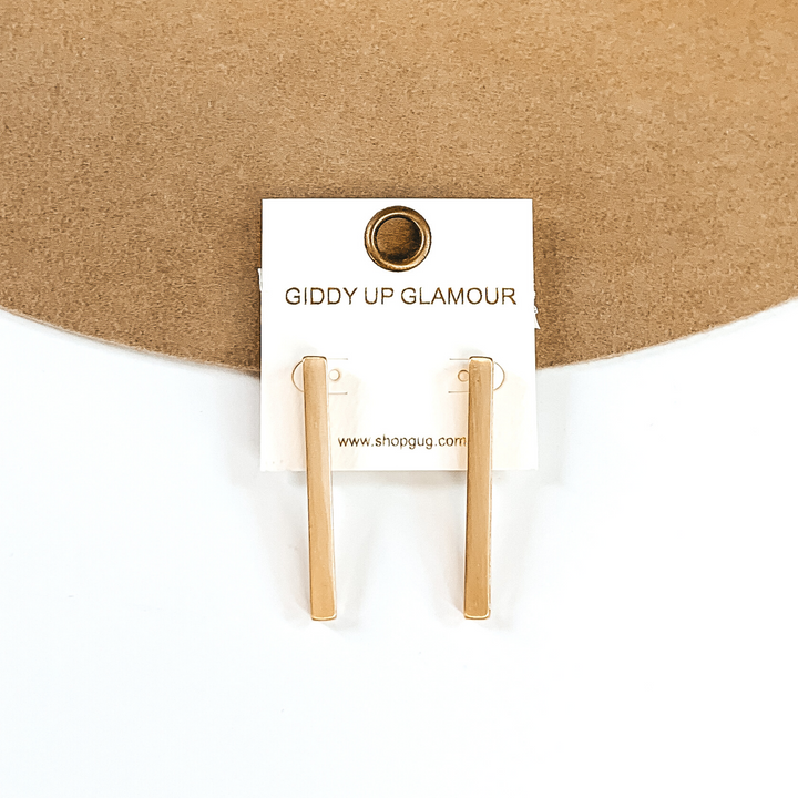 Gold rectangle bar earrings on a white earrings holder. These earrings are pictured on a white and tan background. 