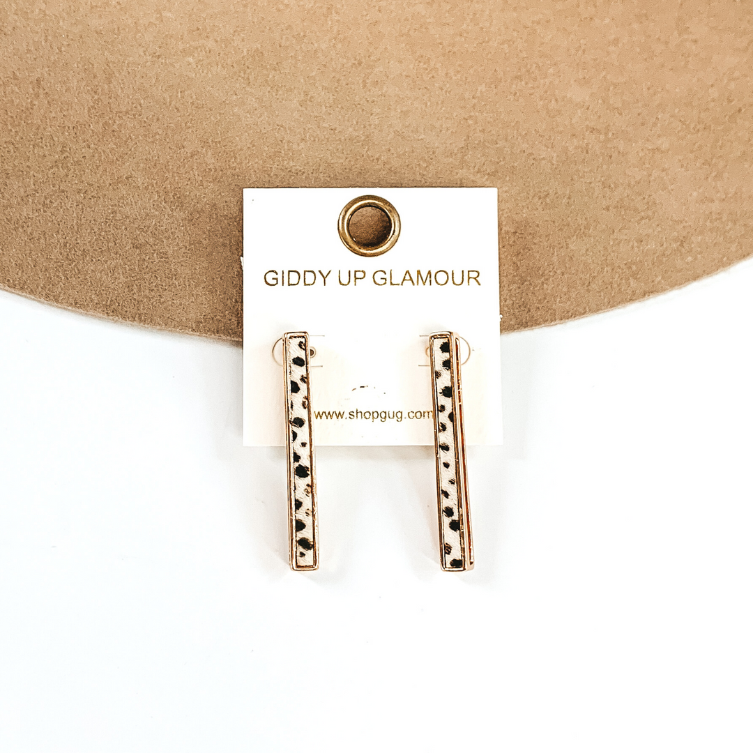 Gold rectangle bar earrings with a white dotted inlay on a white earrings holder. These earrings are pictured on a white and tan background.