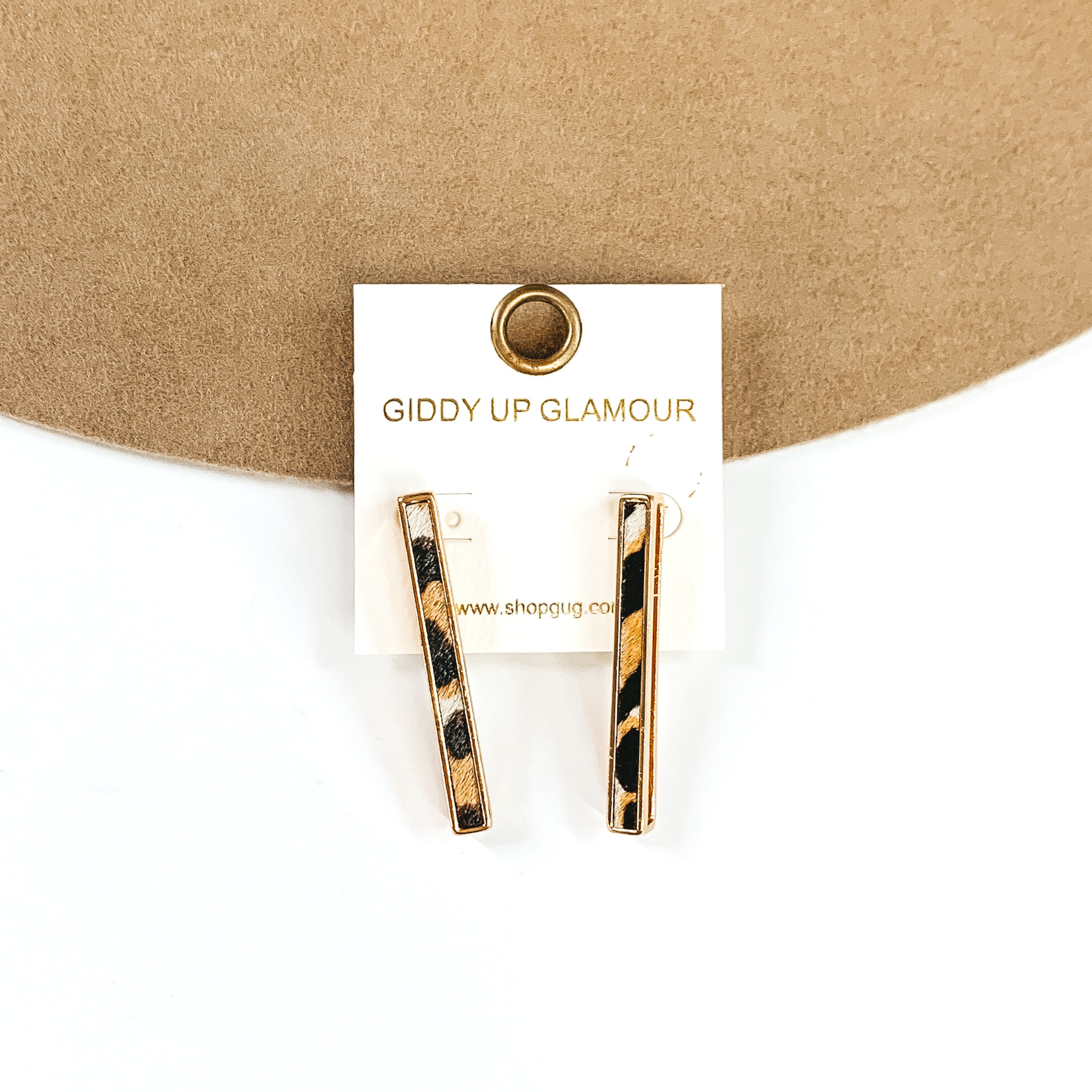 Gold rectangle bar earrings with a white animal print inlay on a white earrings holder. These earrings are pictured on a white and tan background.