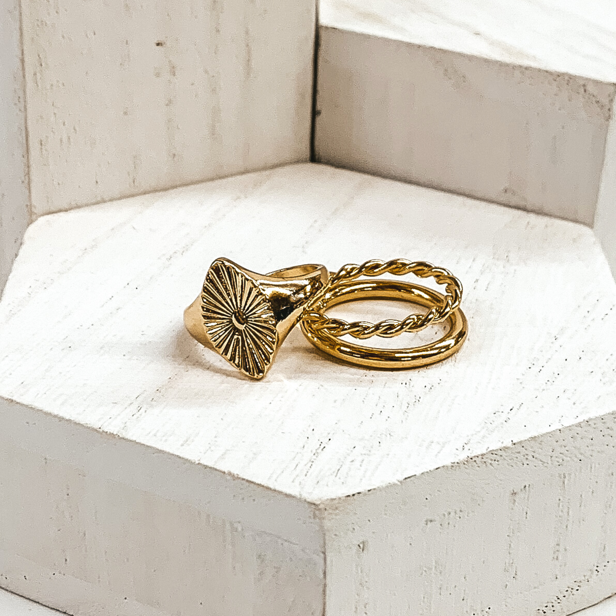 One ring has a marquis shaped pendant with a sunburst design. This ring is laying on a double stacked ring with a plain band connected to a twisted ring. These rings are pictured on white blocks. 