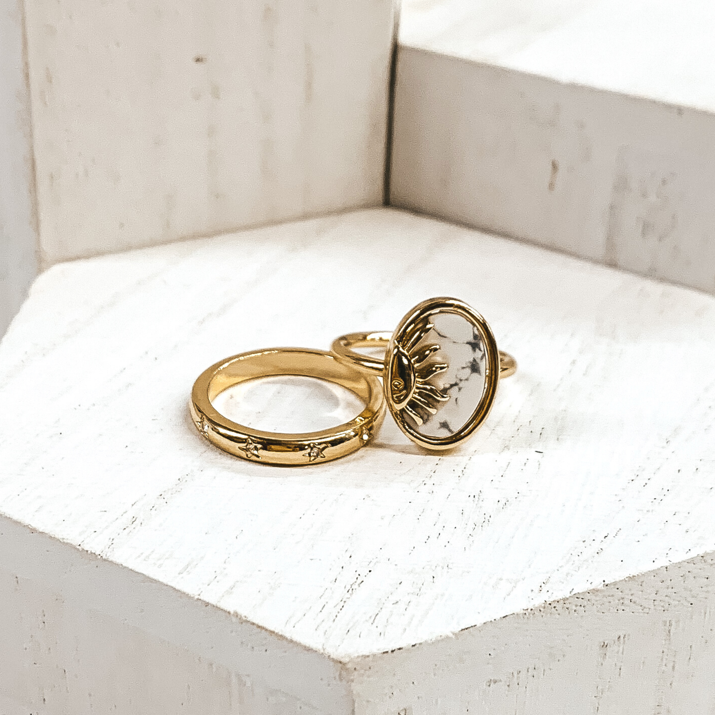 One ring is a plain gold band with tiny, engraved stars with tiny crystals in the center of the stars. The second rings id s plsin gold gand with an oval pendant. The oval pendant is a white stone pendant with a half sun gold charm. These rings are pictured on some white blocks.