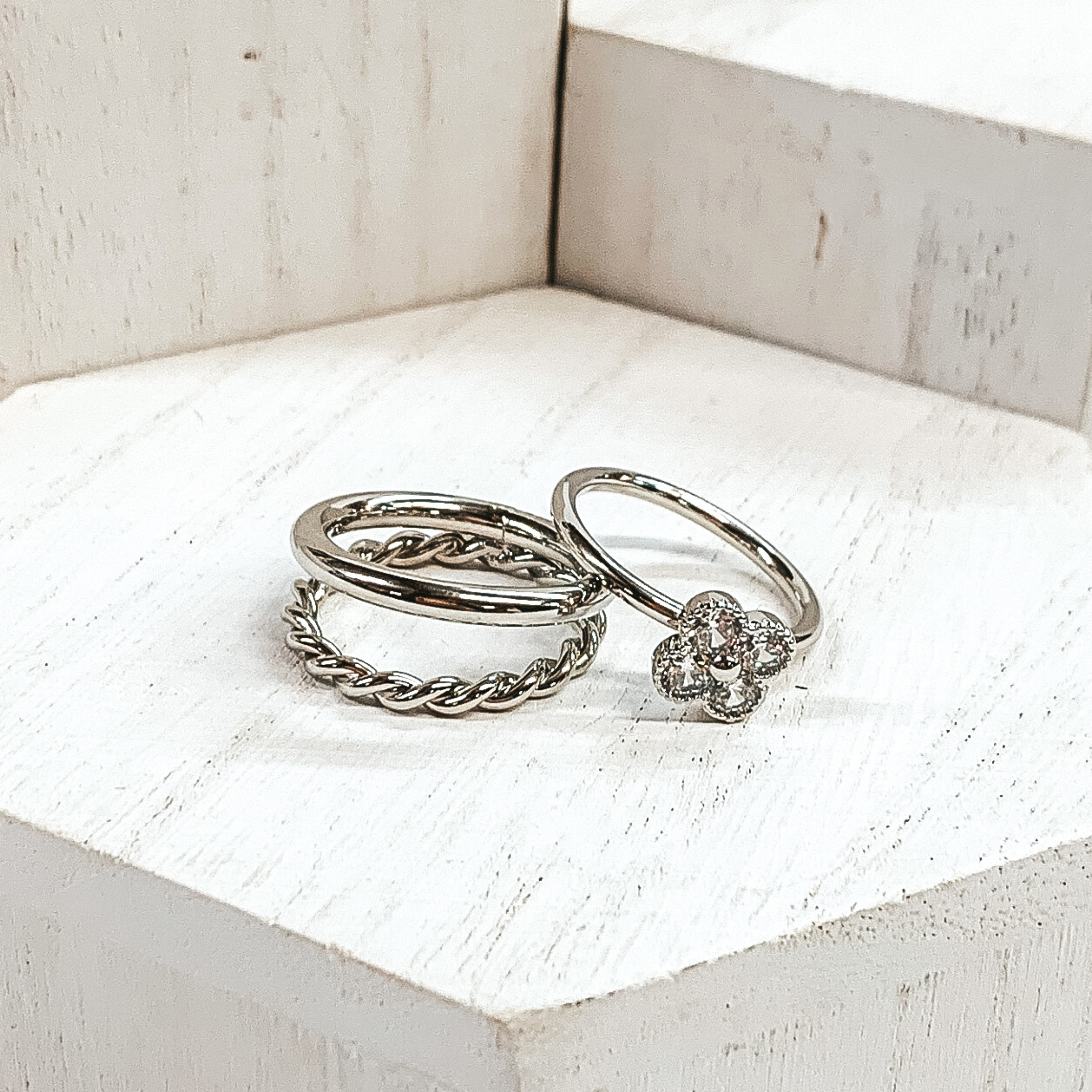 One ring is a silver, double stacked ring with a plain band and twisted band. The second rings is a plain band with a clover pendant that has clear crystals. These rings are pictured on white blocks. 