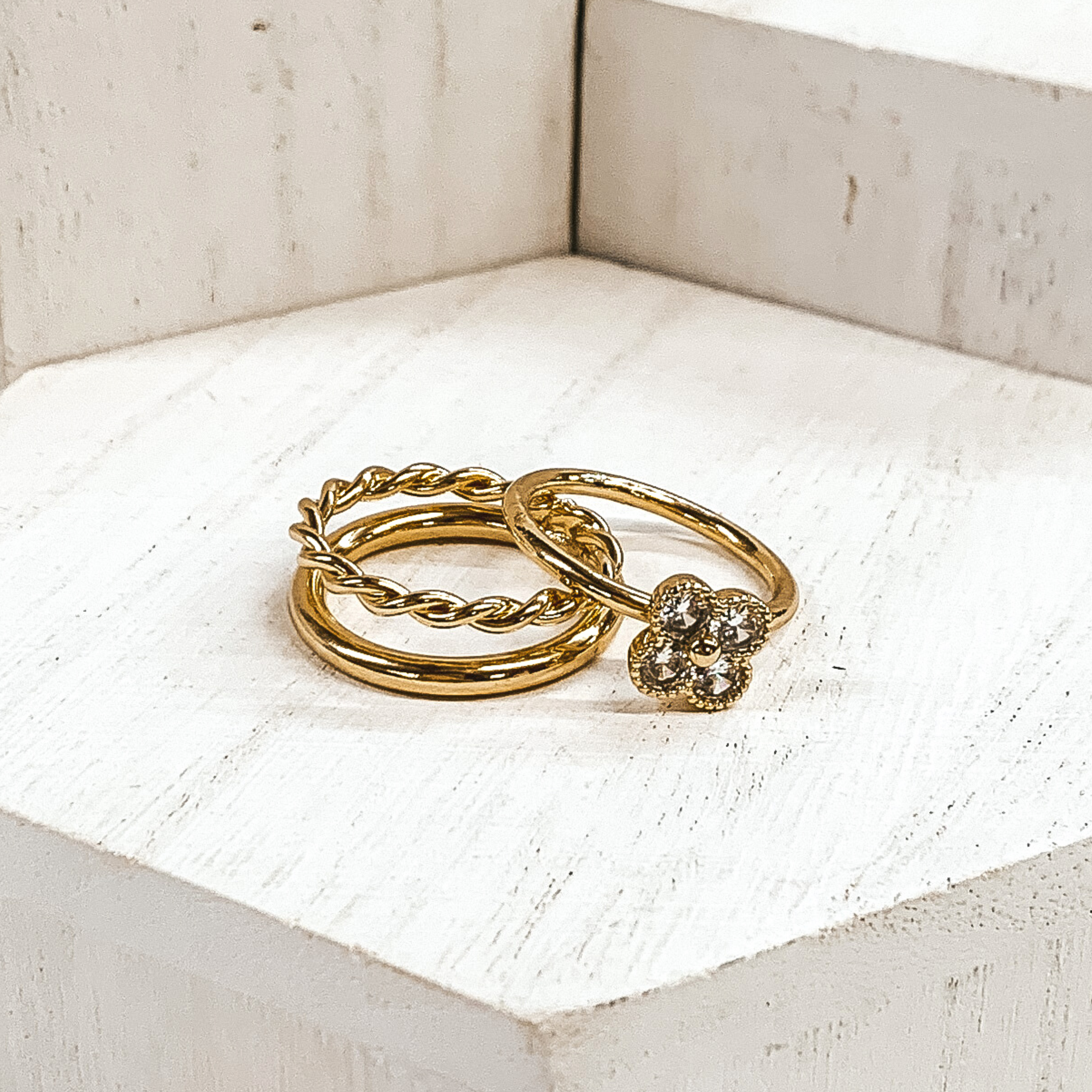 One ring is a gold, double stacked ring with a plain band and twisted band. The second rings is a plain band with a clover pendant that has clear crystals. These rings are pictured on white blocks. 