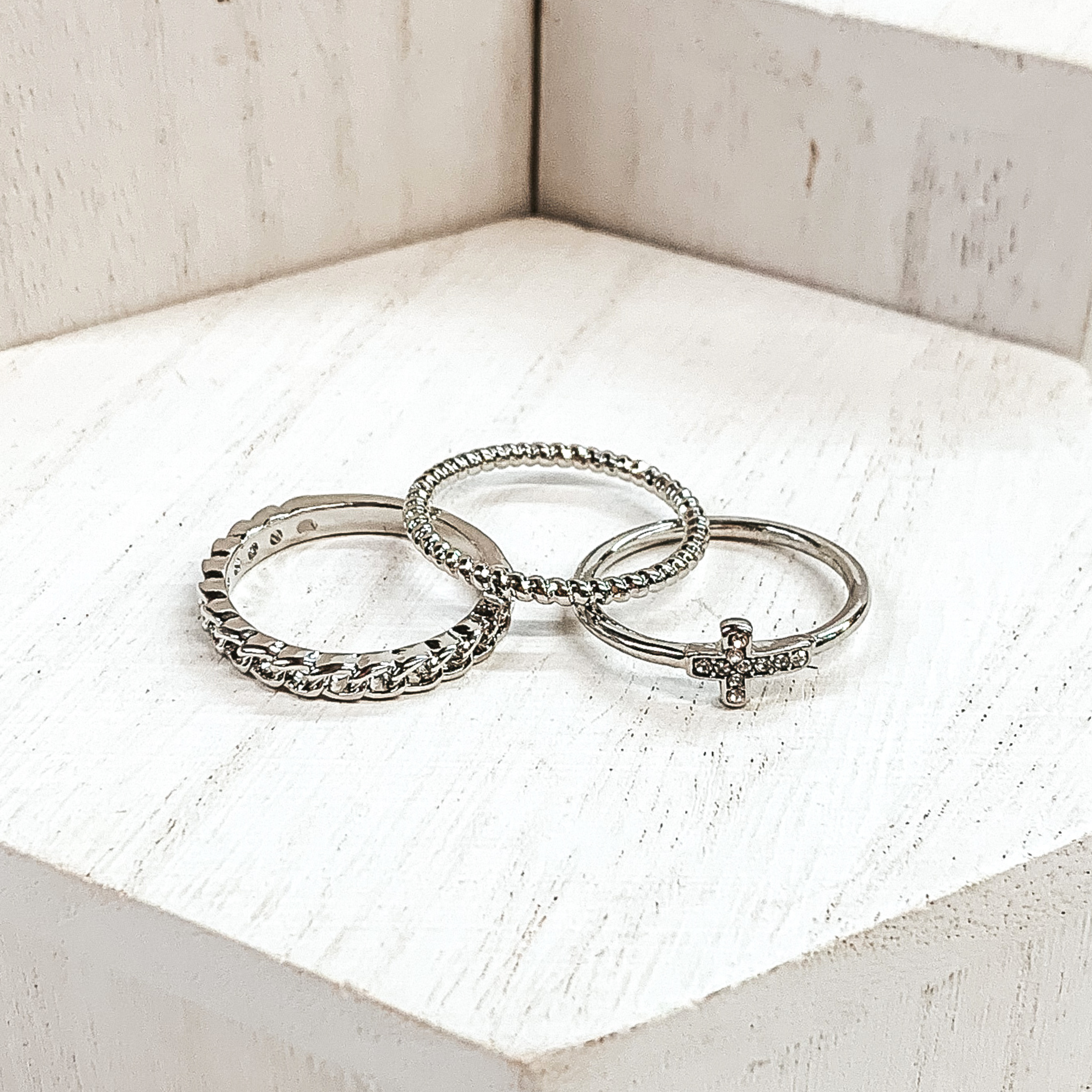 Set of three silver rings. One ring is a twisted ring, another is a half chained ring, and the last ring has a cross pendant that has clear crystals. These rings are pictured on white blocks. 