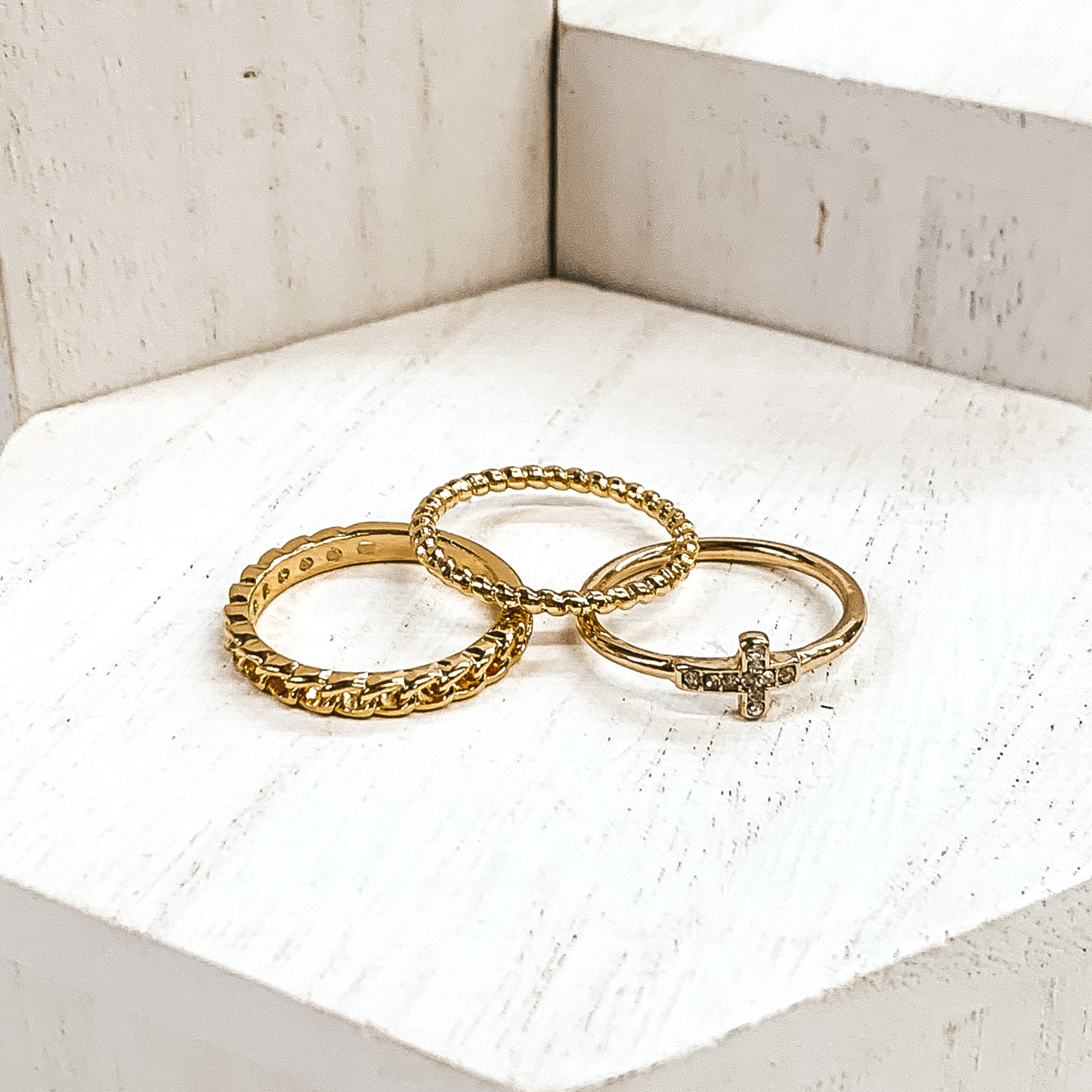 Set of three gold rings. One ring is a twisted ring, another is a half chained ring, and the last ring has a cross pendant that has clear crystals. These rings are pictured on white blocks. 