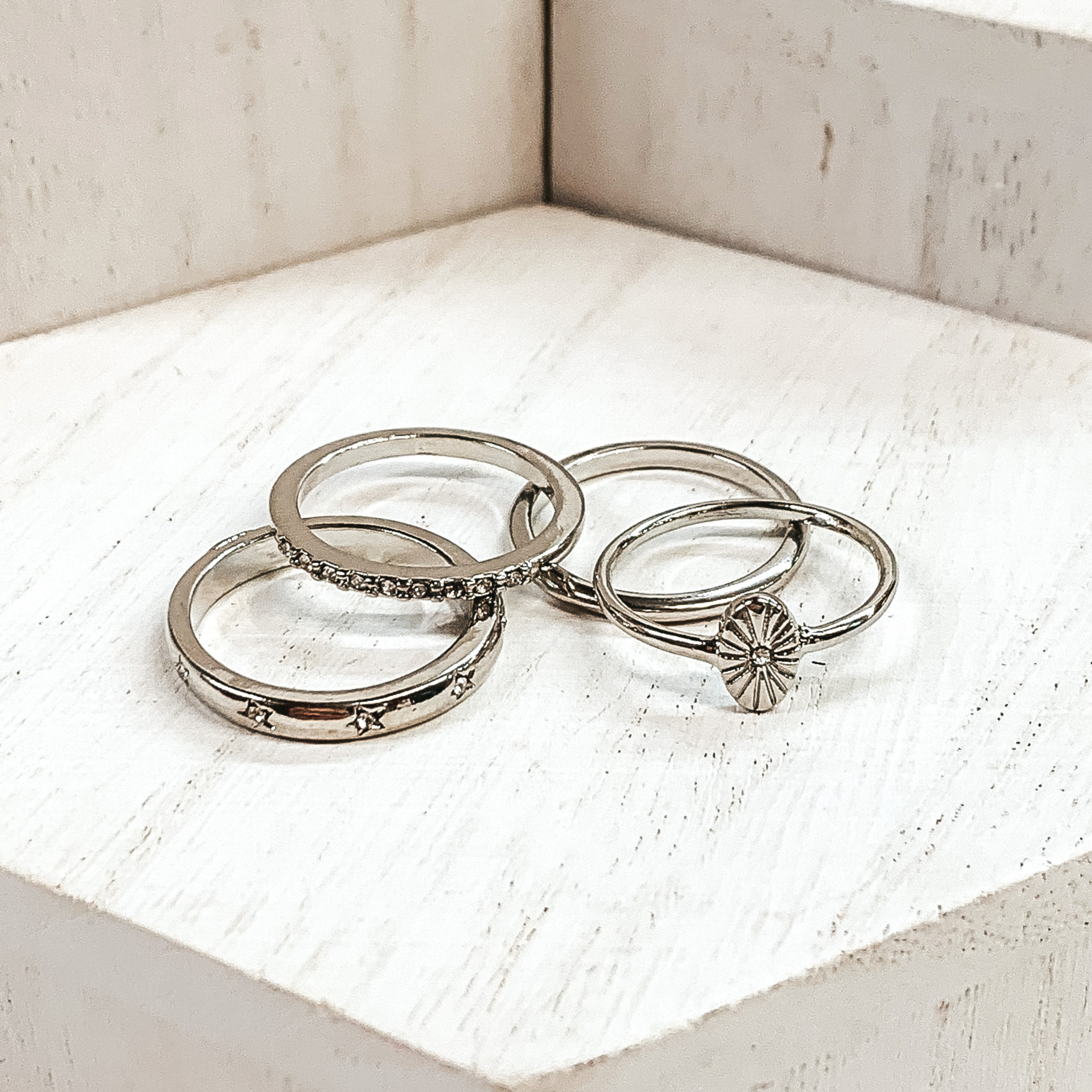 Set of silver rings pictured on white blocks. One ring has an oval pendant, one is a plain band, another ring has clear crystals, and the last ring has tiny engraved stars with tiny clear crystals. 