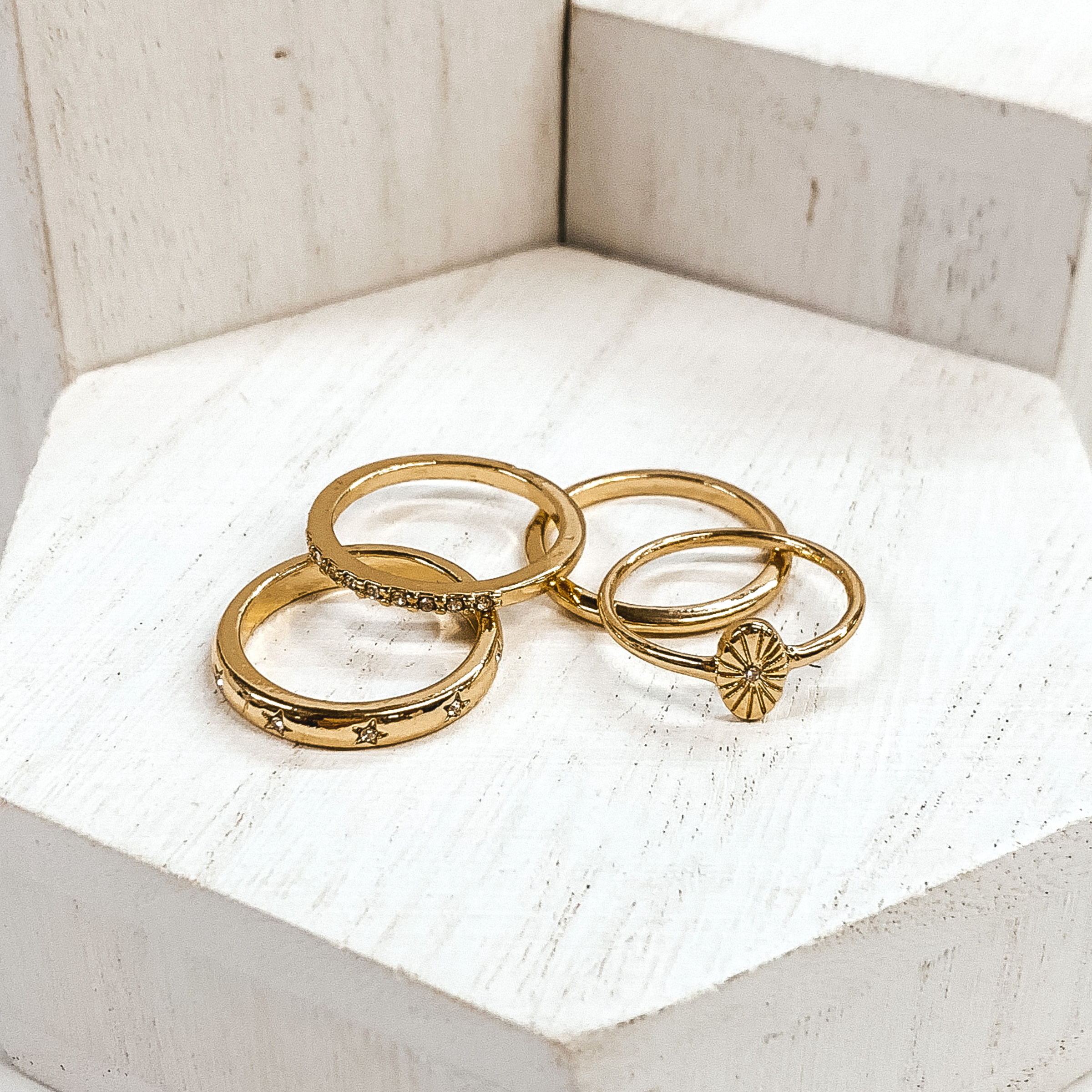 Set of gold rings pictured on white blocks. One ring has an oval pendant, one is a plain band, another ring has clear crystals, and the last ring has tiny engraved stars with tiny clear crystals.