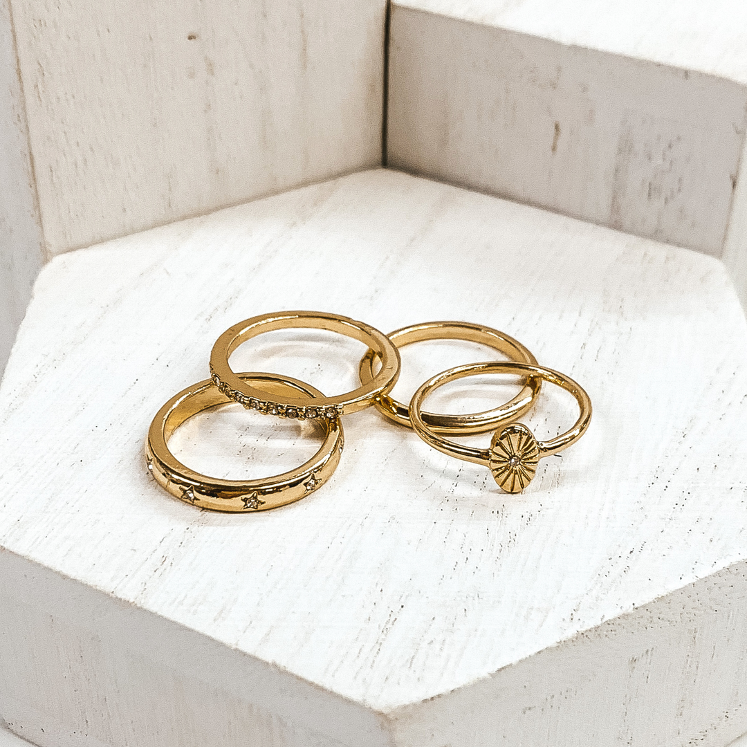 Set of gold rings pictured on white blocks. One ring has an oval pendant, one is a plain band, another ring has clear crystals, and the last ring has tiny engraved stars with tiny clear crystals.