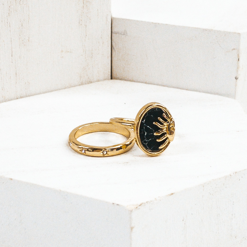 One ring is a plain gold band with tiny, engraved stars with tiny crystals in the center of the stars. The second rings id s plsin gold gand with an oval pendant. The oval pendant is a black stone pendant with a half sun gold charm. These rings are pictured on some white blocks.